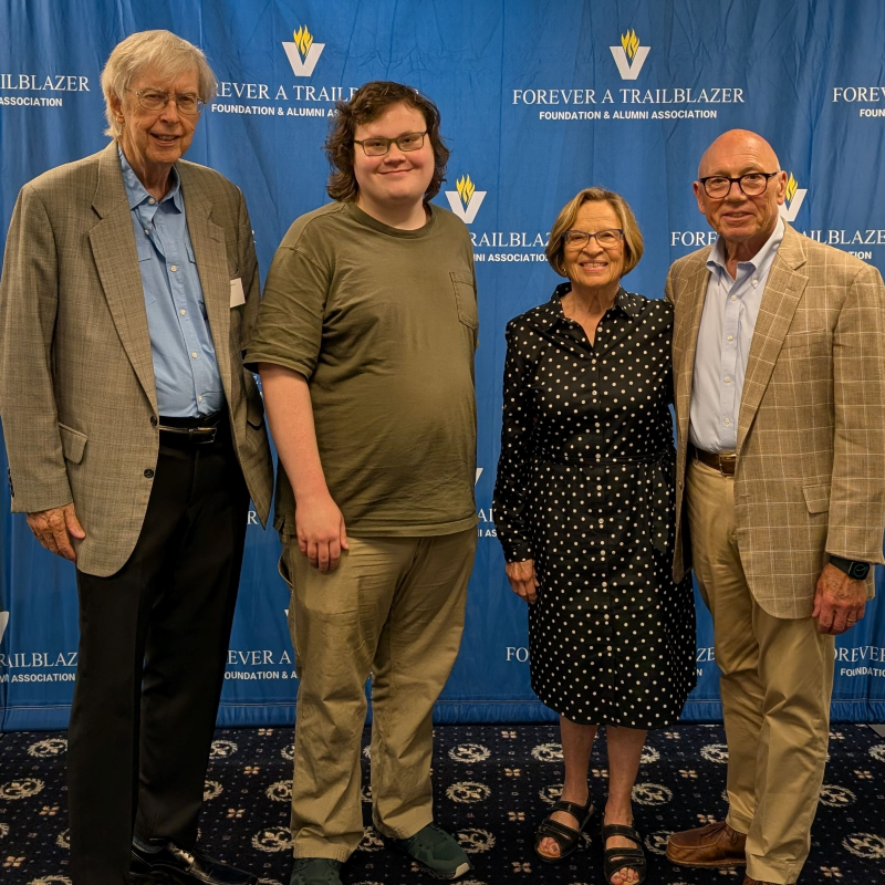 Four people pose together at a formal event in front of a blue “Forever a Trailblazer” foundation backdrop, smiling warmly in business and casual attire.