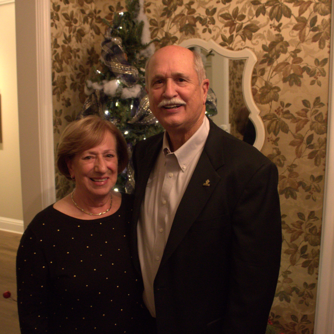 A smiling couple poses beside a lit Christmas tree in a warmly decorated room; he wears a dark suit, she a black dress.
