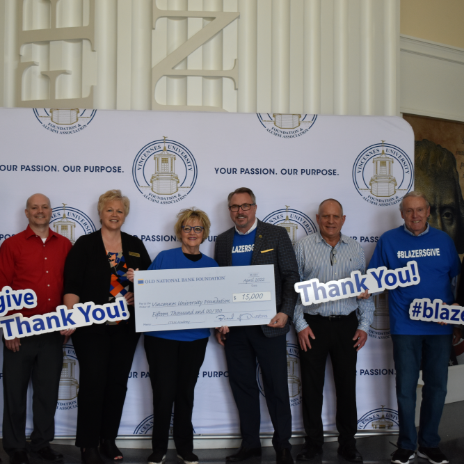 Six smiling adults pose before a branded backdrop, holding oversized blue “Thank You!” signs and a ceremonial check, celebrating a donation or award.