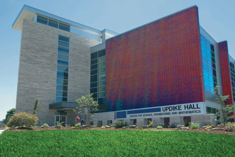 Exterior shot of Updike Hall, a modern academic building that houses the Center for Science, Engineering, and Mathematics. The building features a blend of architectural styles, including a grey stone facade, a vibrant red brick section with colorful hues, and large glass windows reflecting the sky. Landscaping with green manicured grass and small trees complement the building's design. Signage boldly displays "Updike Hall, Center for Science, Engineering and Mathematics."