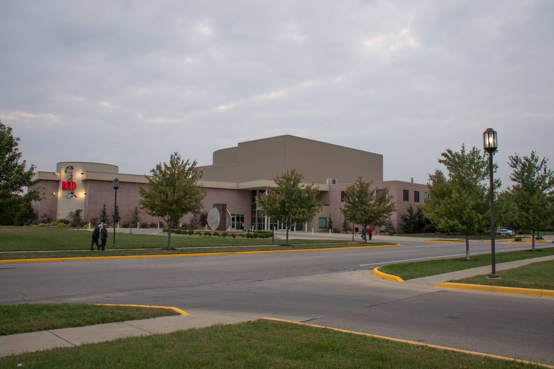 Exterior view of the Red Skelton Performing Arts Center on an overcast day. The building is a mix of light-colored brick and darker metal siding. Trees line the walkways and street around the center. Few people are visible walking towards the entrance. Signage identifies the building with a red logo, featuring a caricature. Yellow curbs highlight the road and sidewalks.