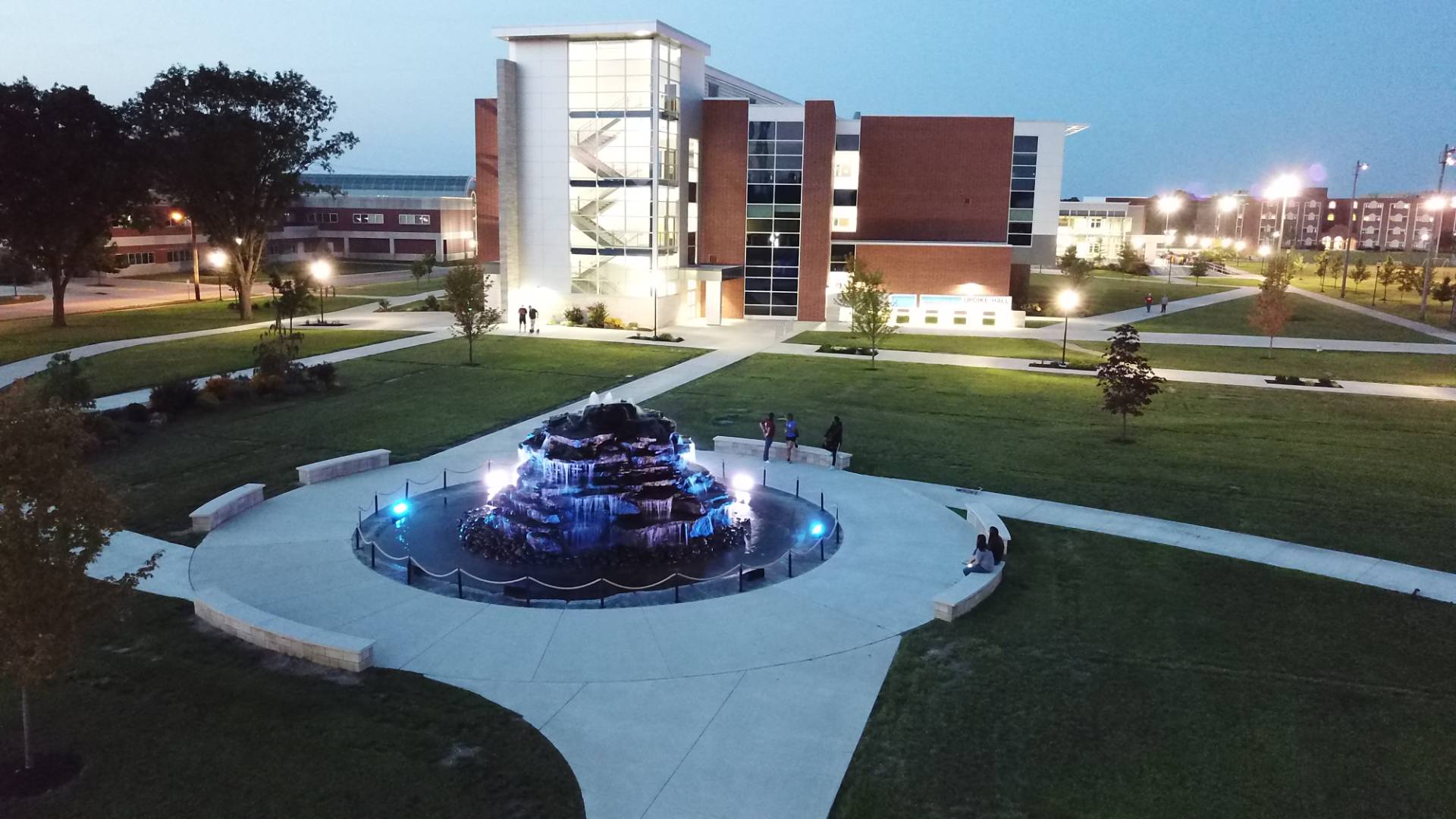 Illuminated campus fountain in front of a modern academic building at dusk.