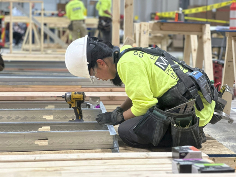 A construction student wearing a hard hat and tool belt kneels to assemble light-gauge steel studs for metal framing, with a cordless drill resting nearby.