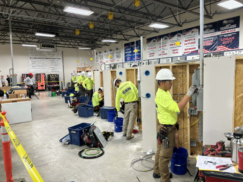 Students wearing hard hats and high-visibility shirts work on electrical wiring and conduit installations at mock wall frames inside a large training facility.