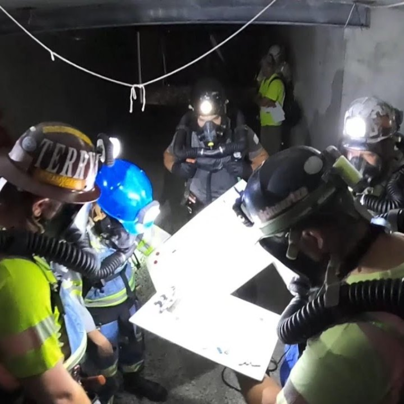 Trainees equipped with mine rescue gear, breathing apparatuses, and headlamps review a schematic map.