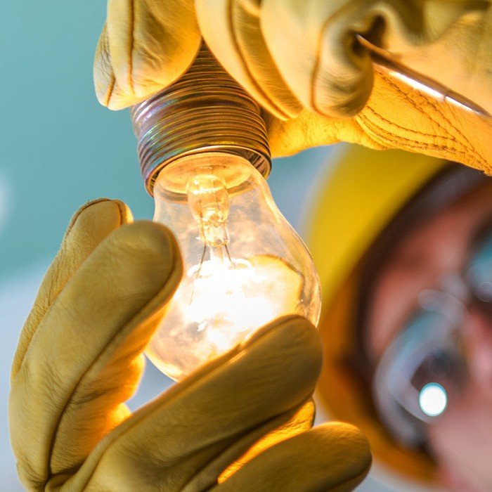 Gloved hands carefully hold a glowing incandescent light bulb against a blurred workshop background.