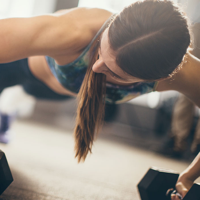 Woman with a ponytail performs a focused dumbbell plank exercise in a bright gym, conveying strength, determination, and an active lifestyle.