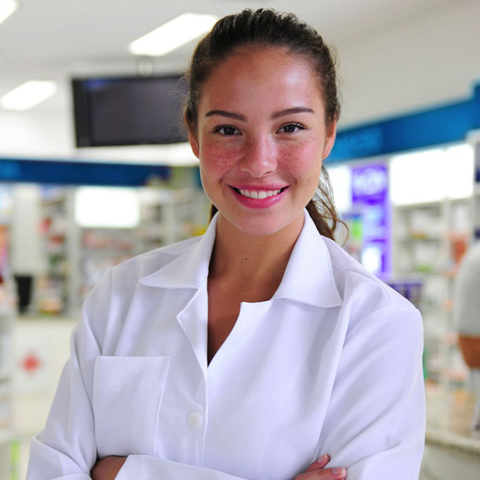 Smiling female pharmacist in a white lab coat stands with folded arms inside a bright pharmacy, conveying warmth, confidence, and professionalism.