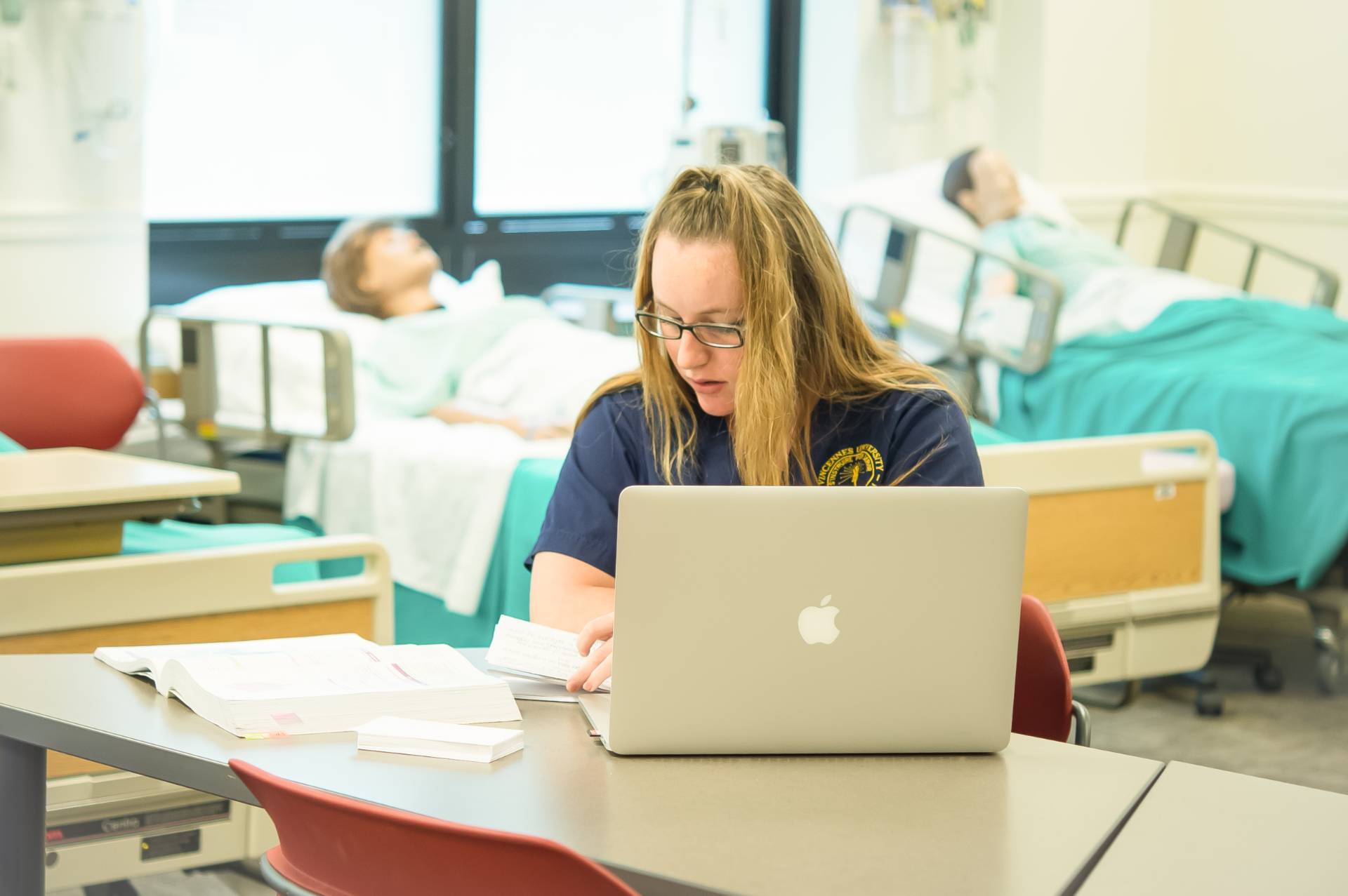 A focused student in a nursing uniform studies at a laptop in a simulation lab with medical dummies in hospital beds, conveying a learning environment.