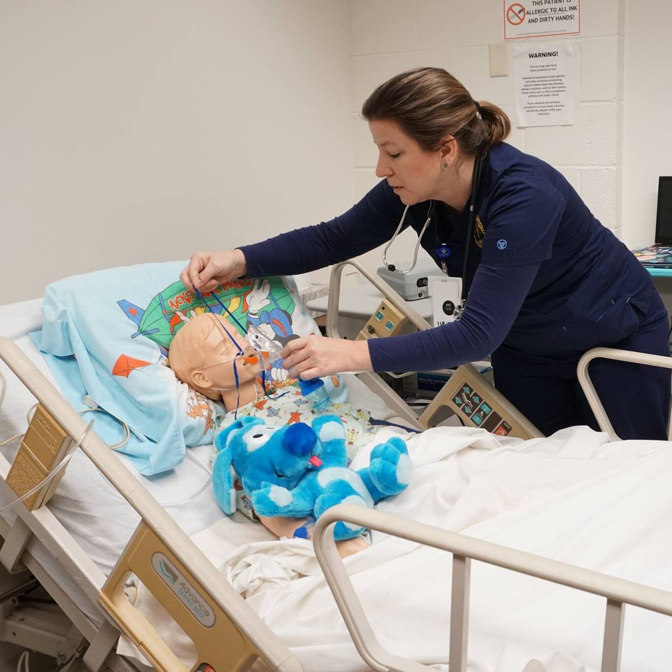 In a brightly lit hospital room, a nurse wearing a dark blue scrub suit adjusts an oxygen mask on a child mannequin lying in a hospital bed. The mannequin is dressed in pajamas and has a light-colored wig. Beside the mannequin is a plush toy of Blue from Blue's Clues. The hospital bed has a light blue Mickey Mouse-themed pillowcase. Medical equipment is visible in the background. On the wall is a sign that reads, "Warning! THIS PATIENT IS ALLERGIC TO ALL INK AND DIRTY HANDS!".