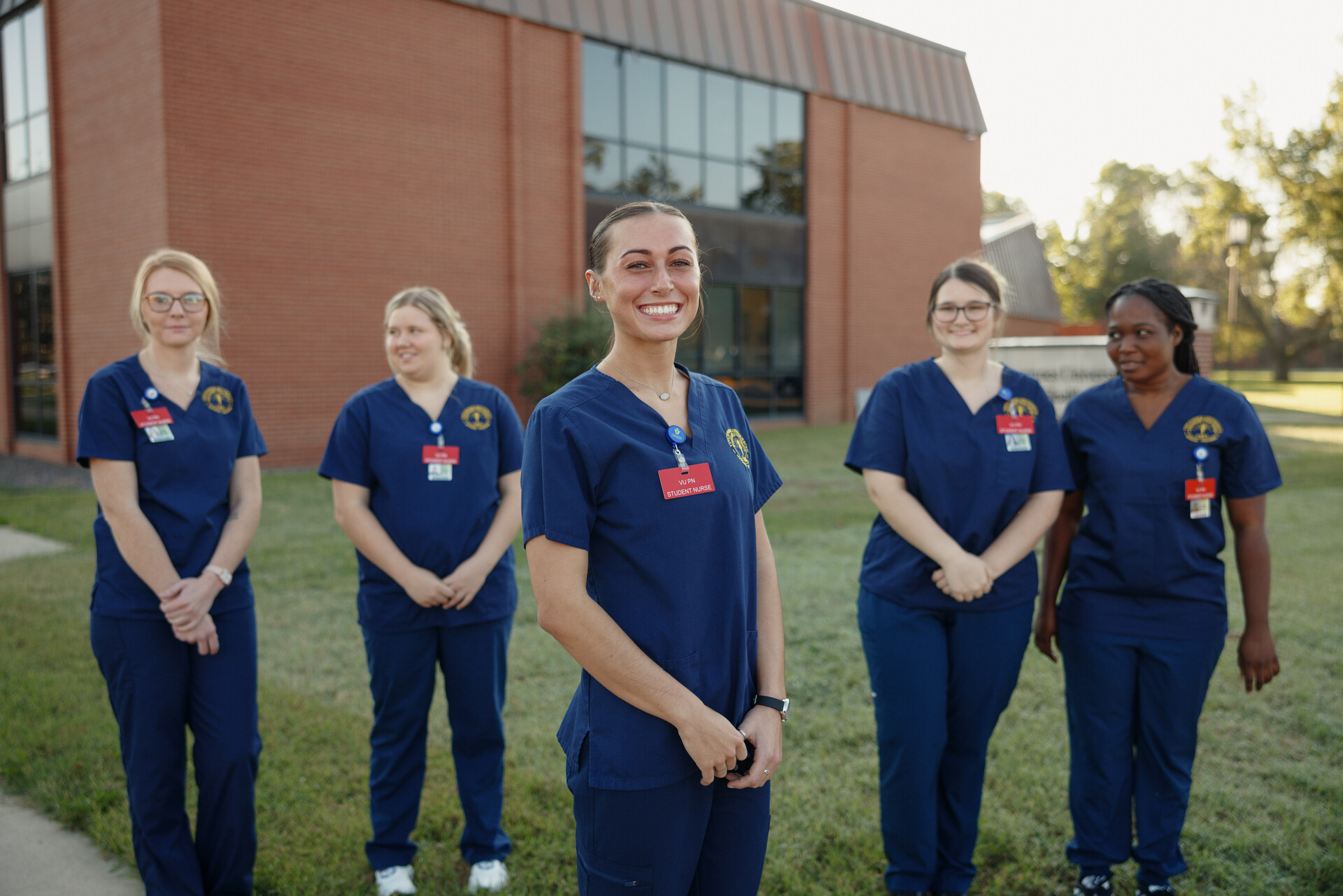 A group of five nursing students stands outdoors in navy scrubs, focusing on teamwork and solidarity, with a red brick building behind them.