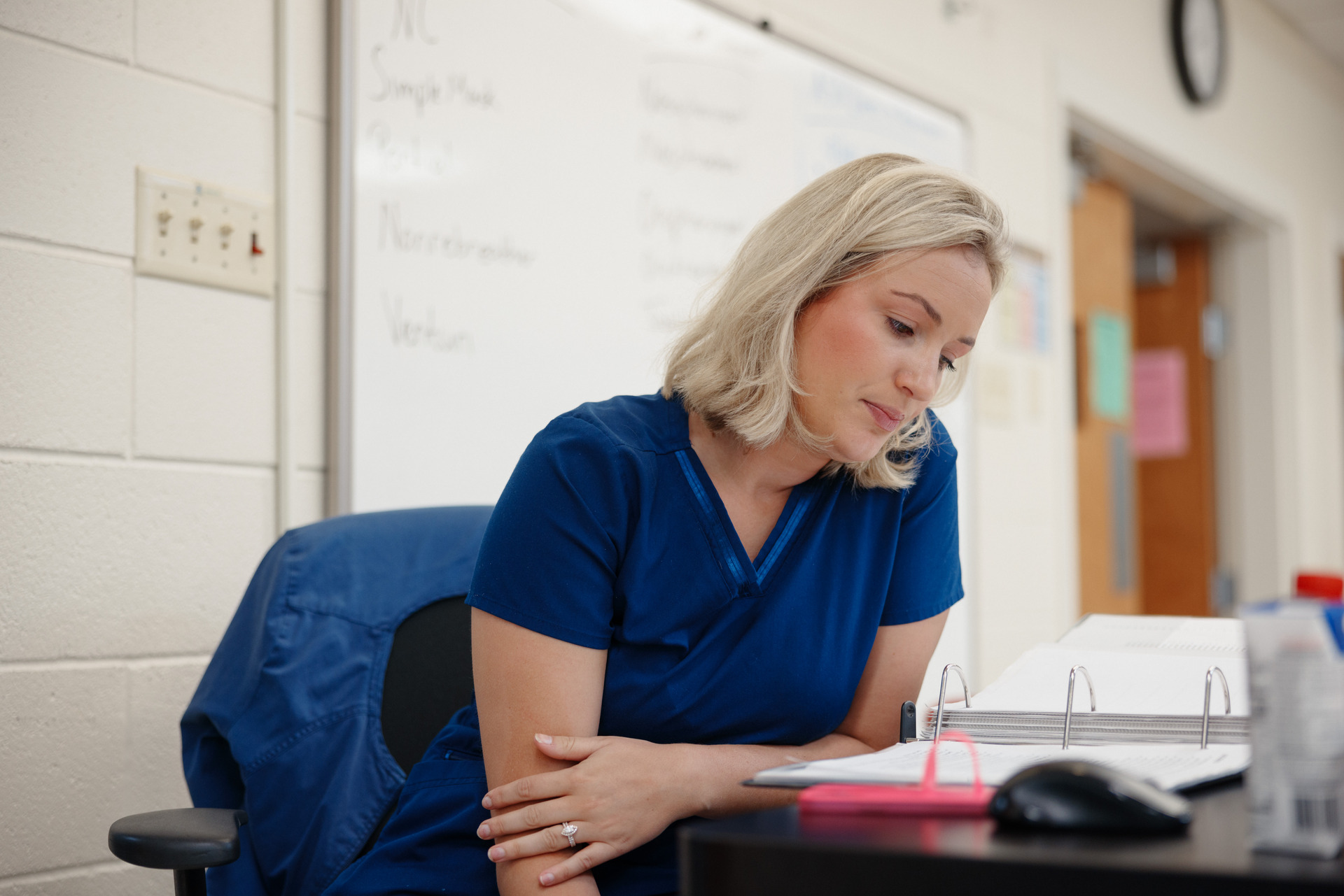 Nursing student in blue scrubs studying at a desk with an open binder in a classroom setting, focused on exam preparation or coursework.
