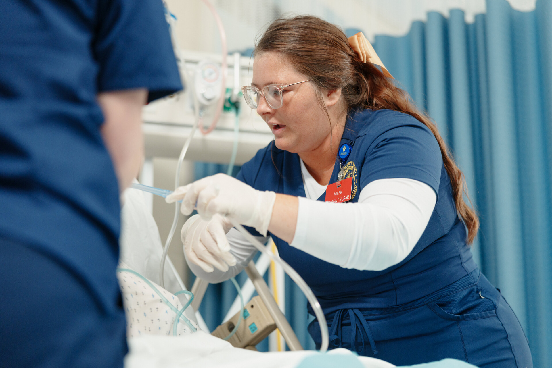 A nursing student in blue scrubs and gloves tends to a patient in a hospital bed. She appears focused and attentive, adjusting medical tubing equipment.