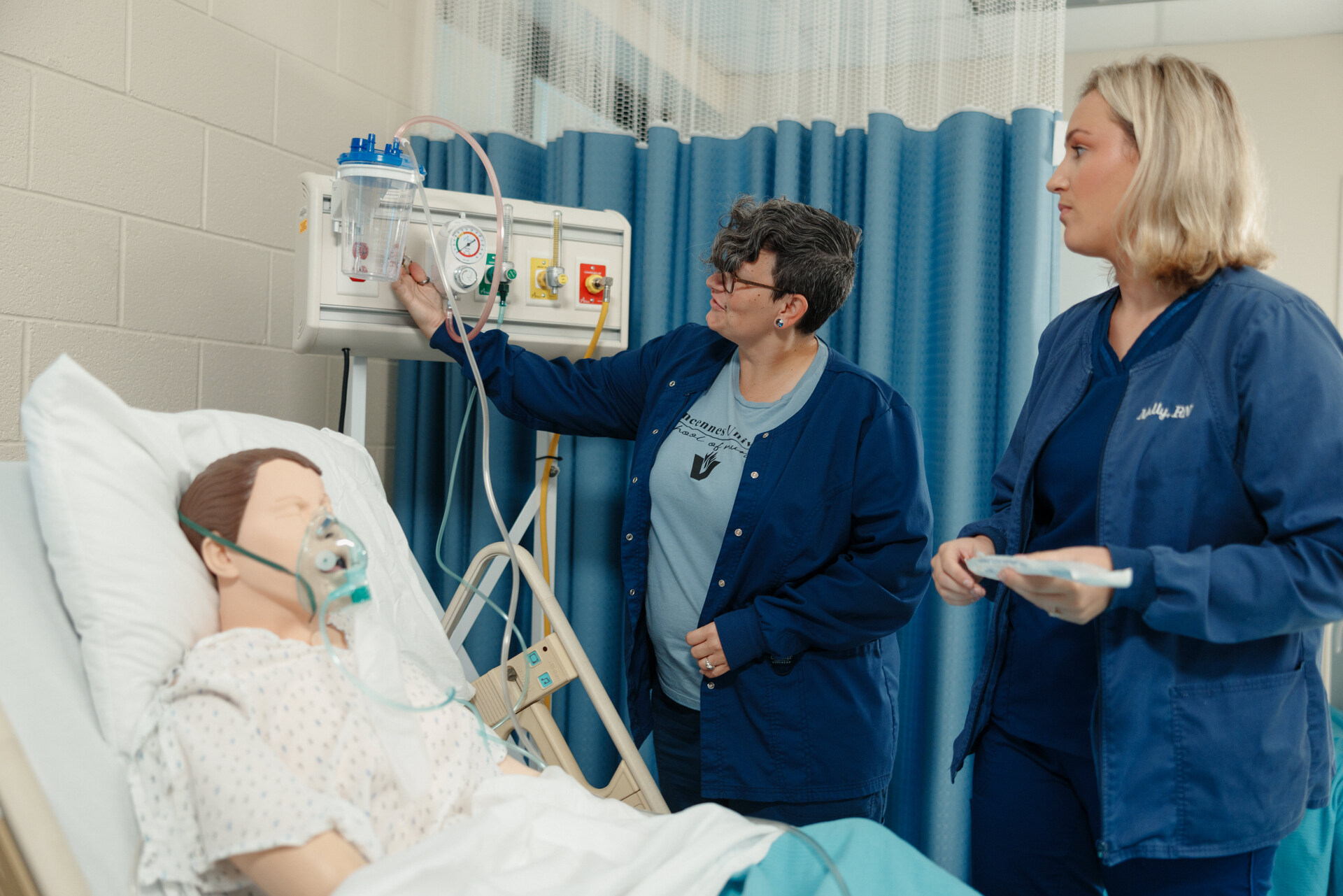 Two nursing students are attending to a patient simulation in a hospital setting. One nurse is adjusting equipment on the wall, while the other is holding a clipboard. The patient, represented by a mannequin wearing a hospital gown and an oxygen mask, is lying in a hospital bed. The background features blue privacy curtains and medical equipment, creating a realistic training environment.