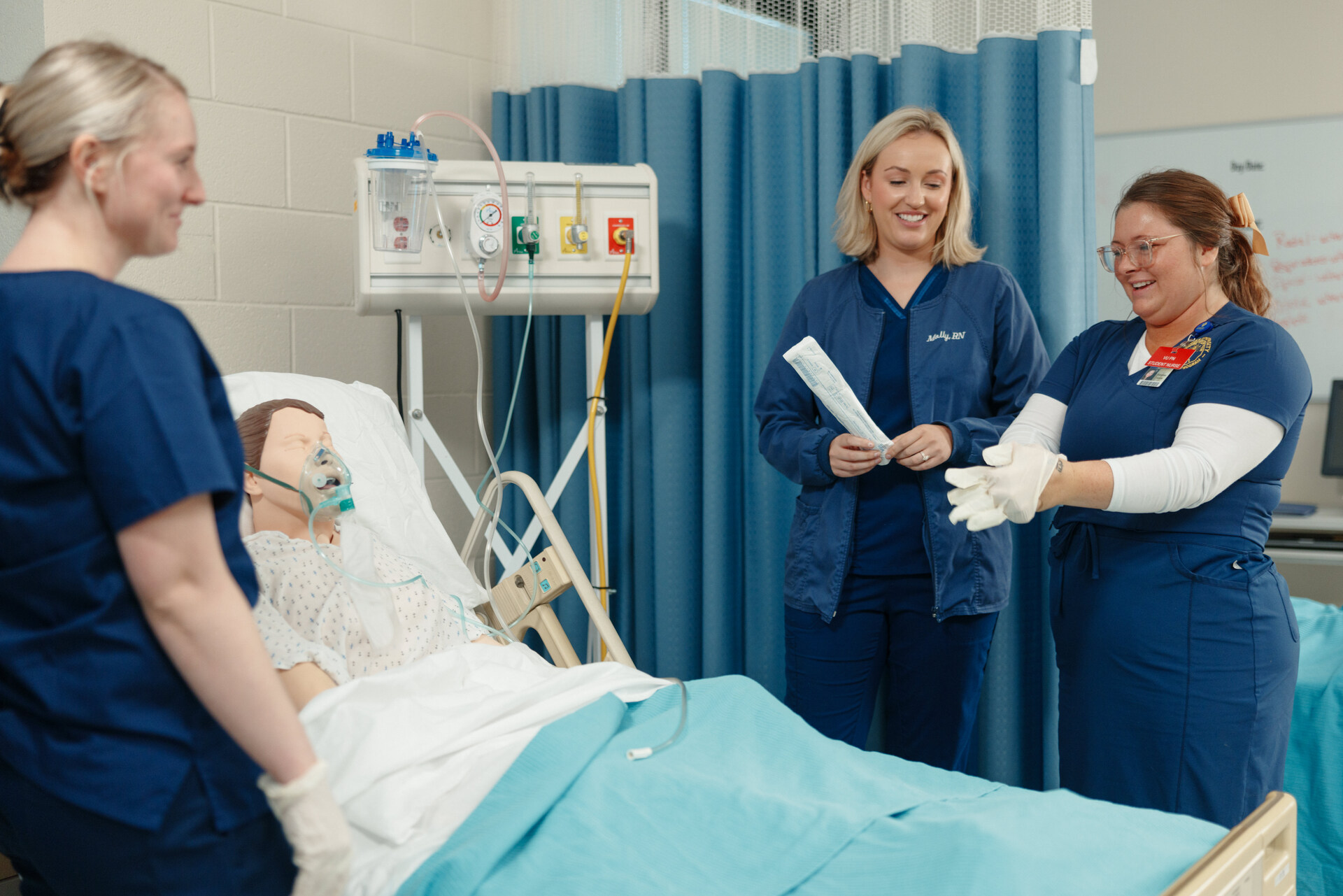 Nursing students practice a medical scenario using a hospital bed mannequin, while an instructor supervises and demonstrates proper technique in a clinical lab setting.
