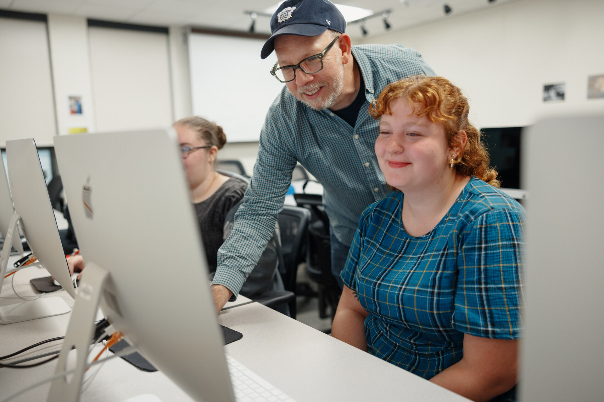 A middle-aged male instructor with glasses and wearing a blue baseball cap, stands behind a young female student as she sits at a desk with an iMac computer. They are both looking at the screen. A second student sits at a computer in the background. The scene takes place in a classroom.
