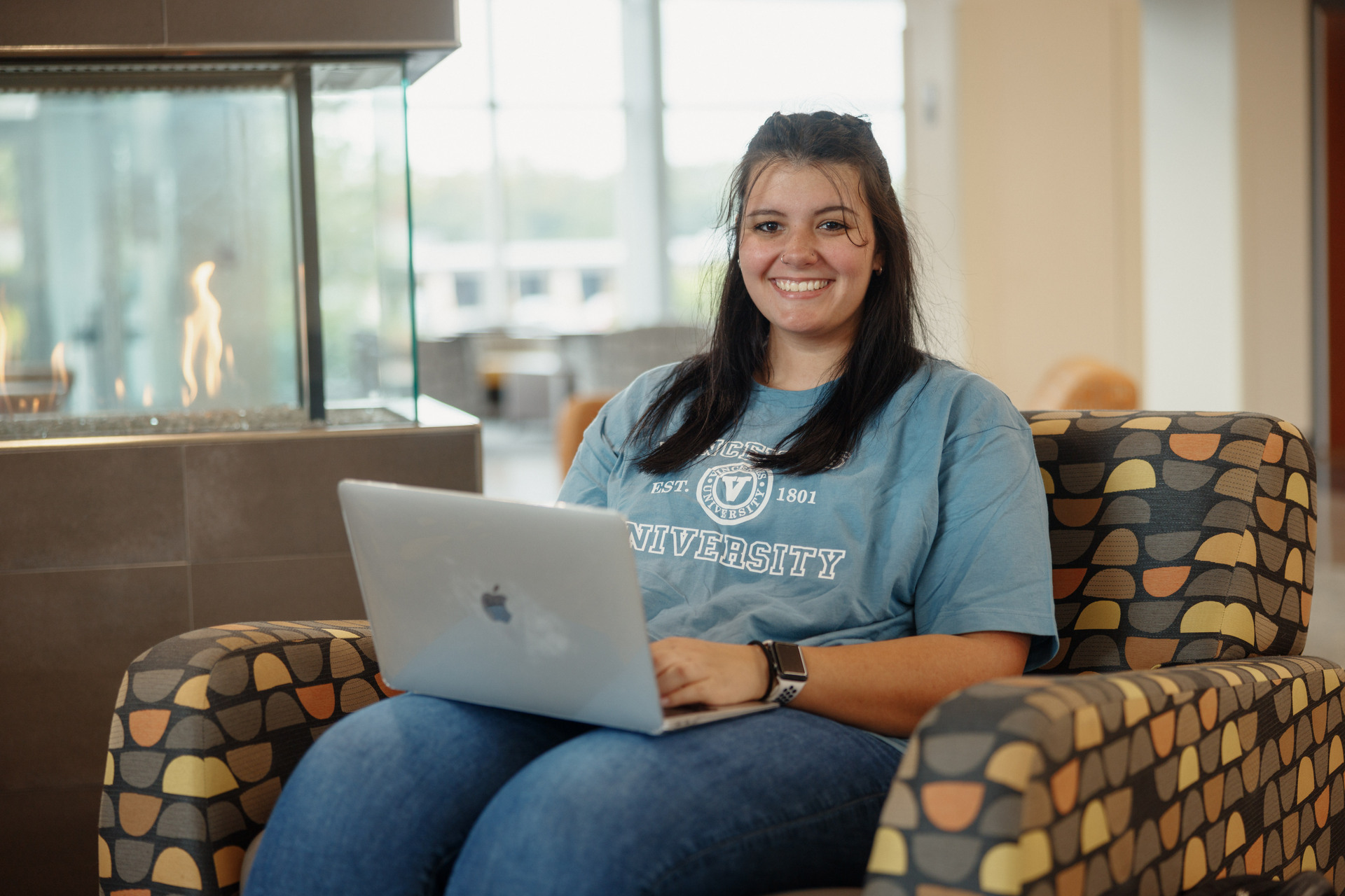 A smiling dark-haired young woman sits in a patterned armchair while using a laptop. She is wearing a blue t-shirt with a college logo and jeans. A modern fireplace with visible flames is visible in the background, along with a brightly lit interior space.