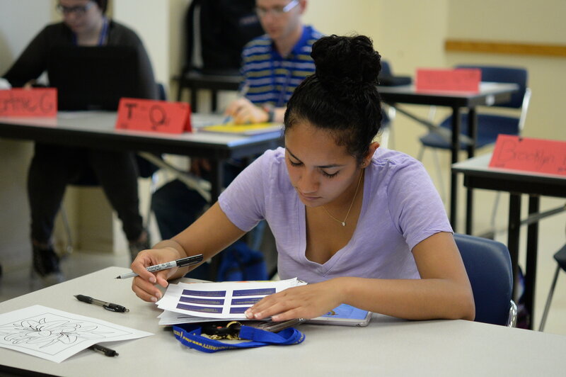 A young woman with her hair in a bun is seated at a desk in a classroom, focused on completing a worksheet. She holds a black marker in her right hand and is looking down at the paper. Other students and desks with name cards on them are visible in the background. The classroom appears to be set up for an activity or exam.