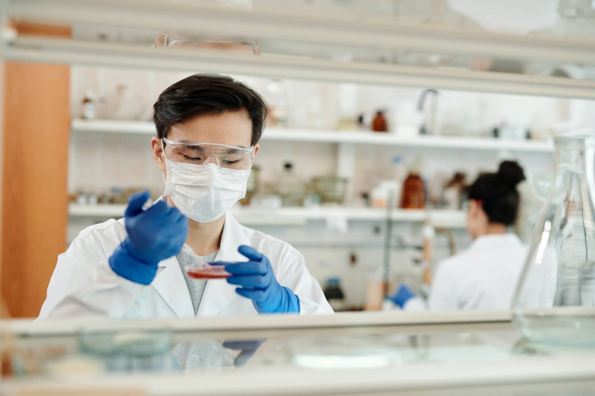 Scientist in protective gear and gloves examines a petri dish in a lab. Another scientist works in the background. The scene conveys focus and precision.