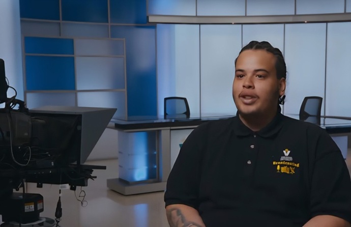 A person is seated in a modern TV studio with blue accents, wearing a black polo shirt. The background shows empty chairs and a camera setup. The atmosphere is professional and calm.