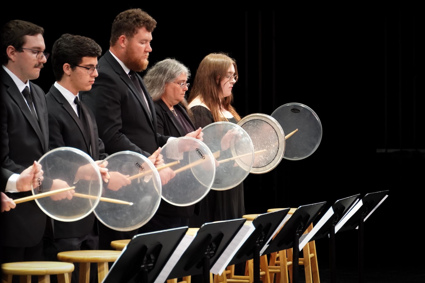 Five musicians in formal attire hold clear drumheads with mallets, standing next to stools and music stands, focused in a performance setting.