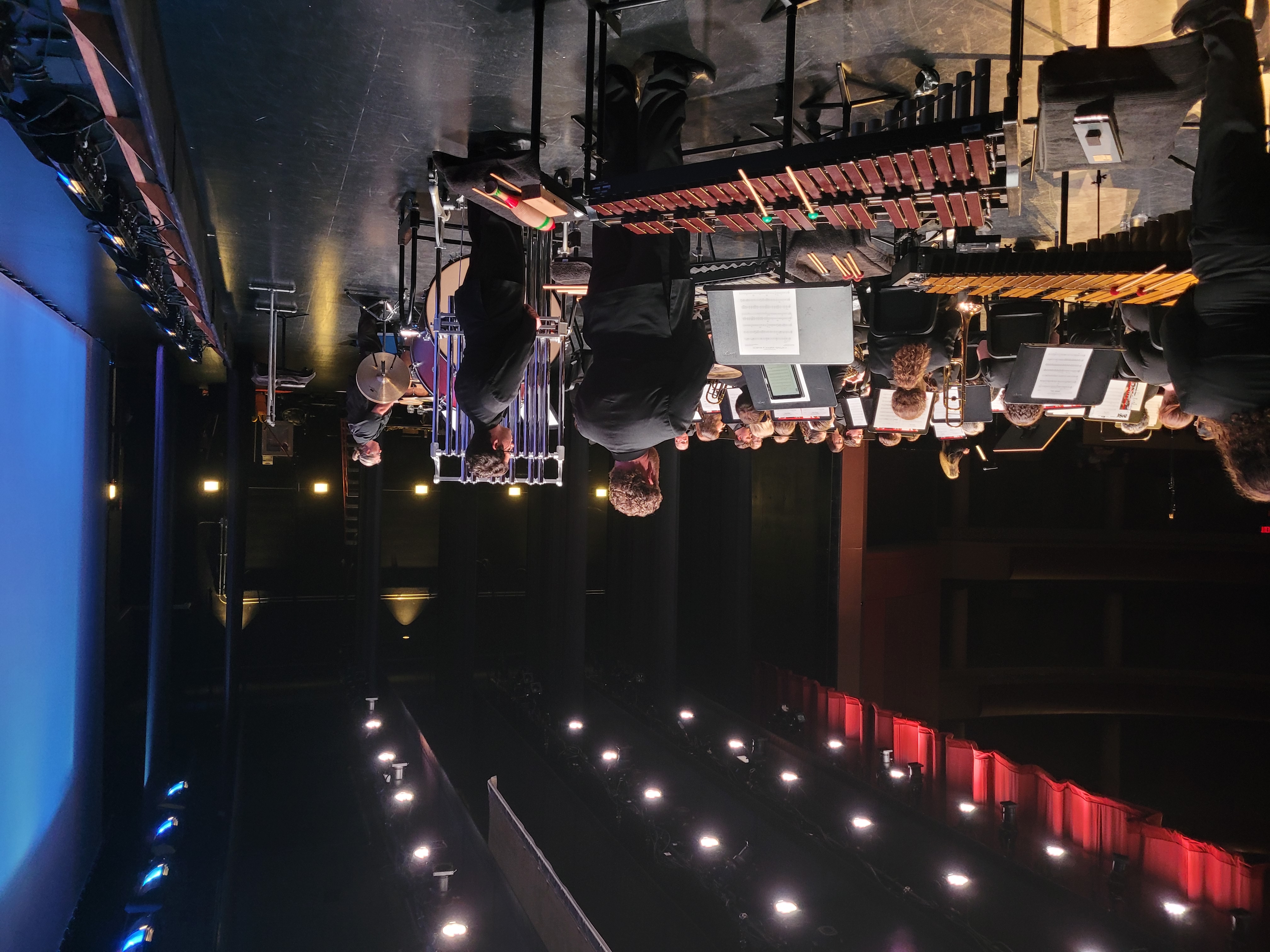 A symphony orchestra on stage, musicians in black formal wear. The conductor stands at the front. Percussion instruments are visible, with warm, dramatic lighting.