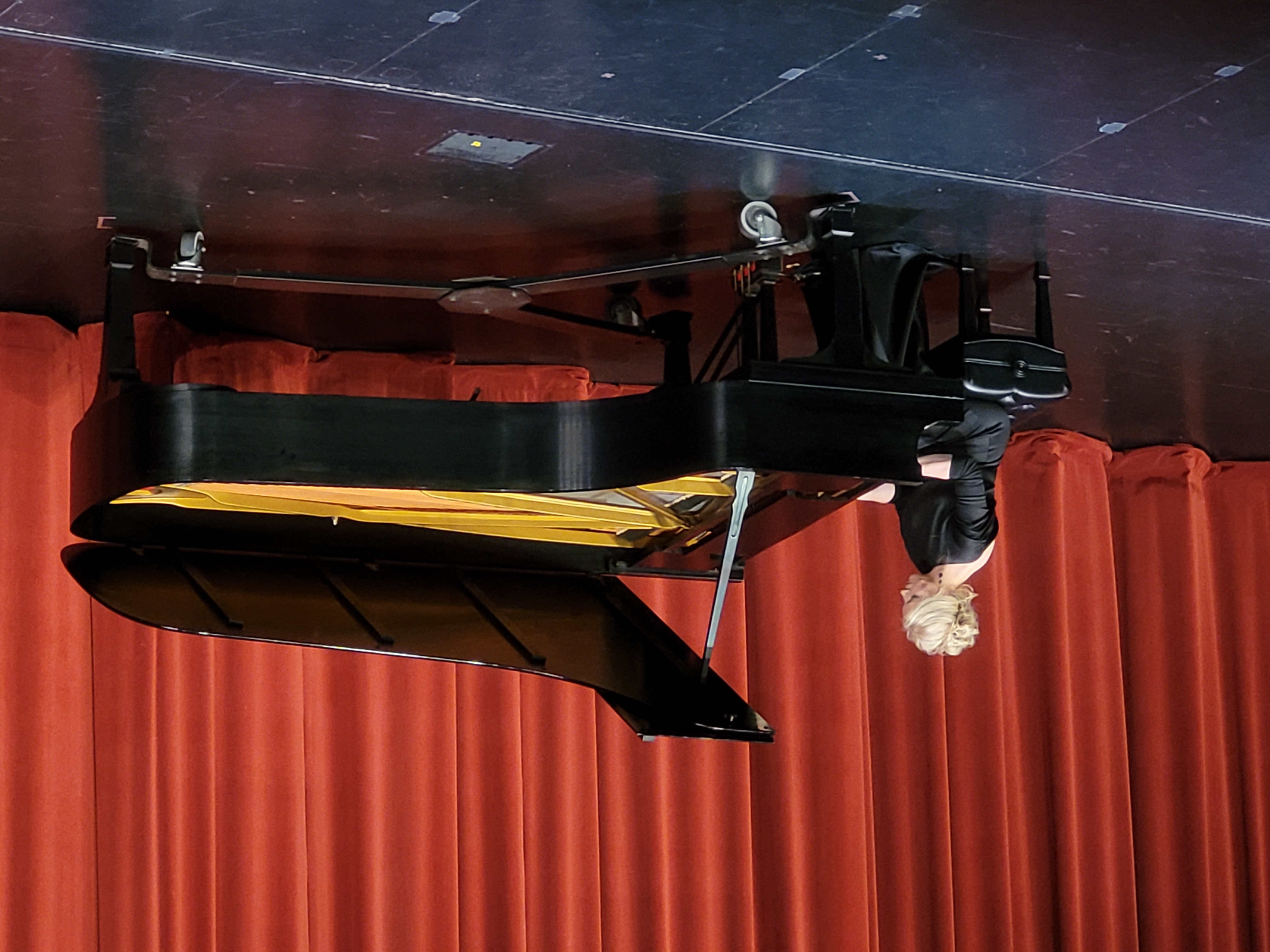 A woman in a black gown plays a grand piano on stage. The background features red curtains, creating an elegant and focused atmosphere.