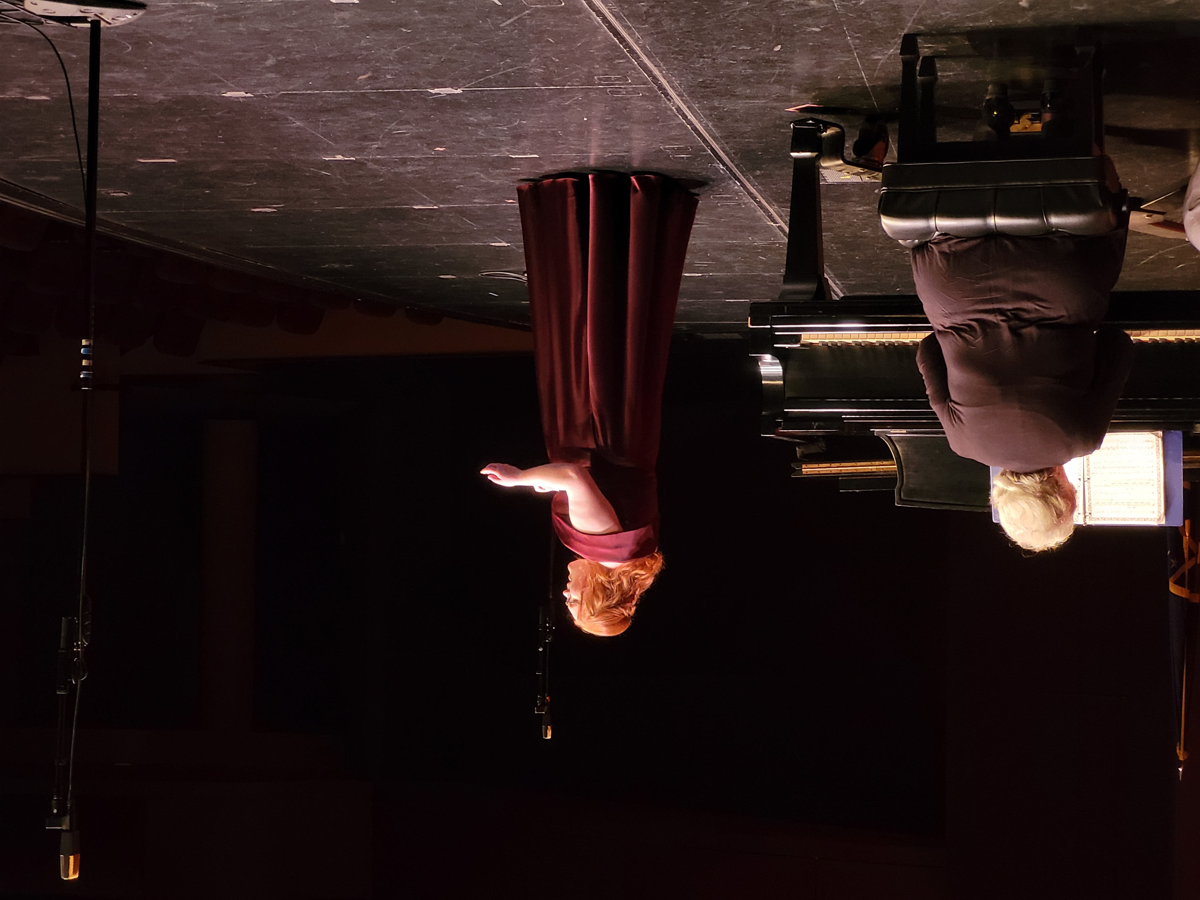 A woman in a flowing maroon gown sings passionately on stage, while an older woman plays the piano. The scene is dimly lit, creating an intimate and emotional atmosphere.