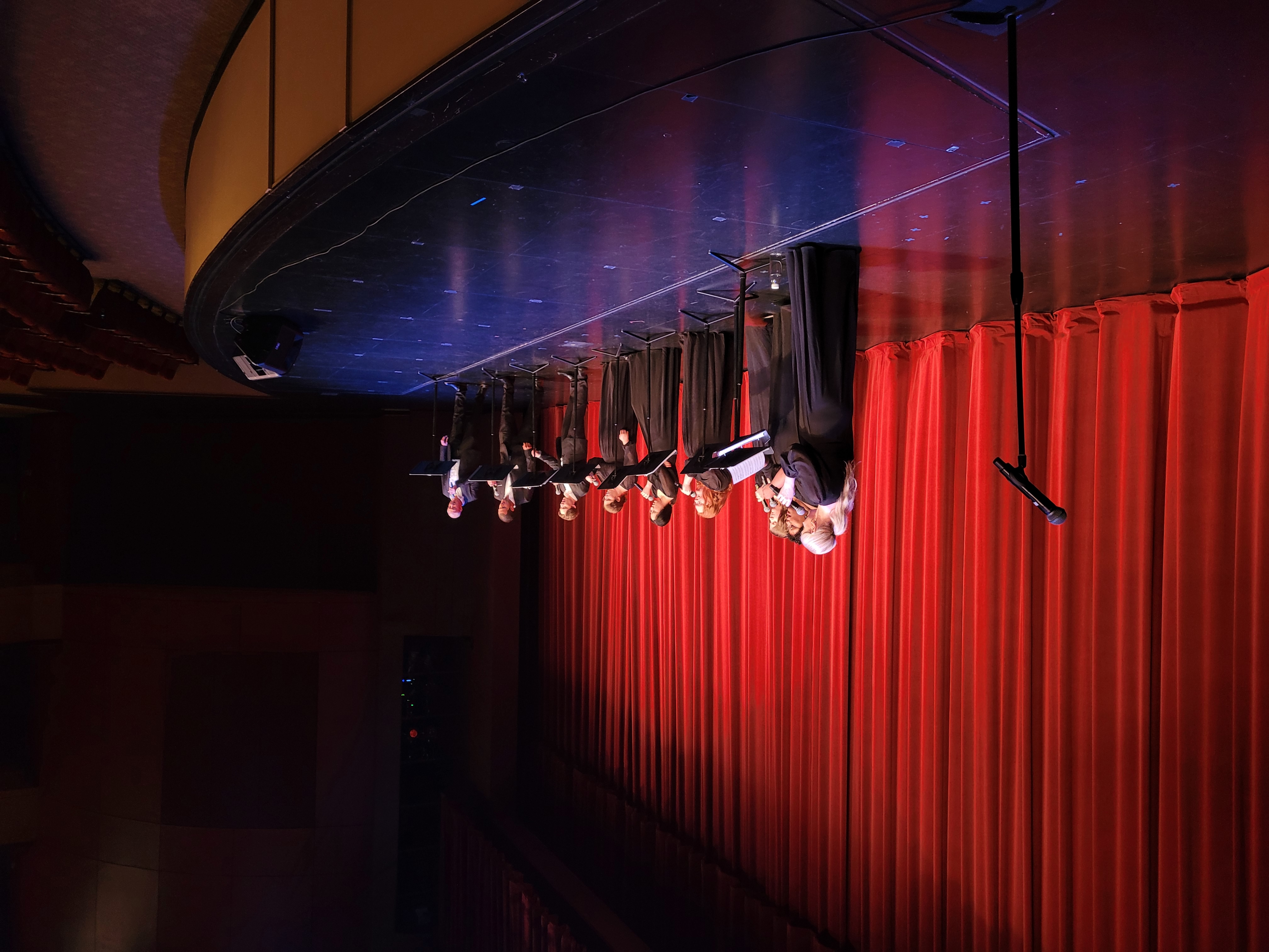 A group of singers in formal black attire performs on a brightly lit stage, with red curtains as the backdrop, exuding a sense of elegance and focus.