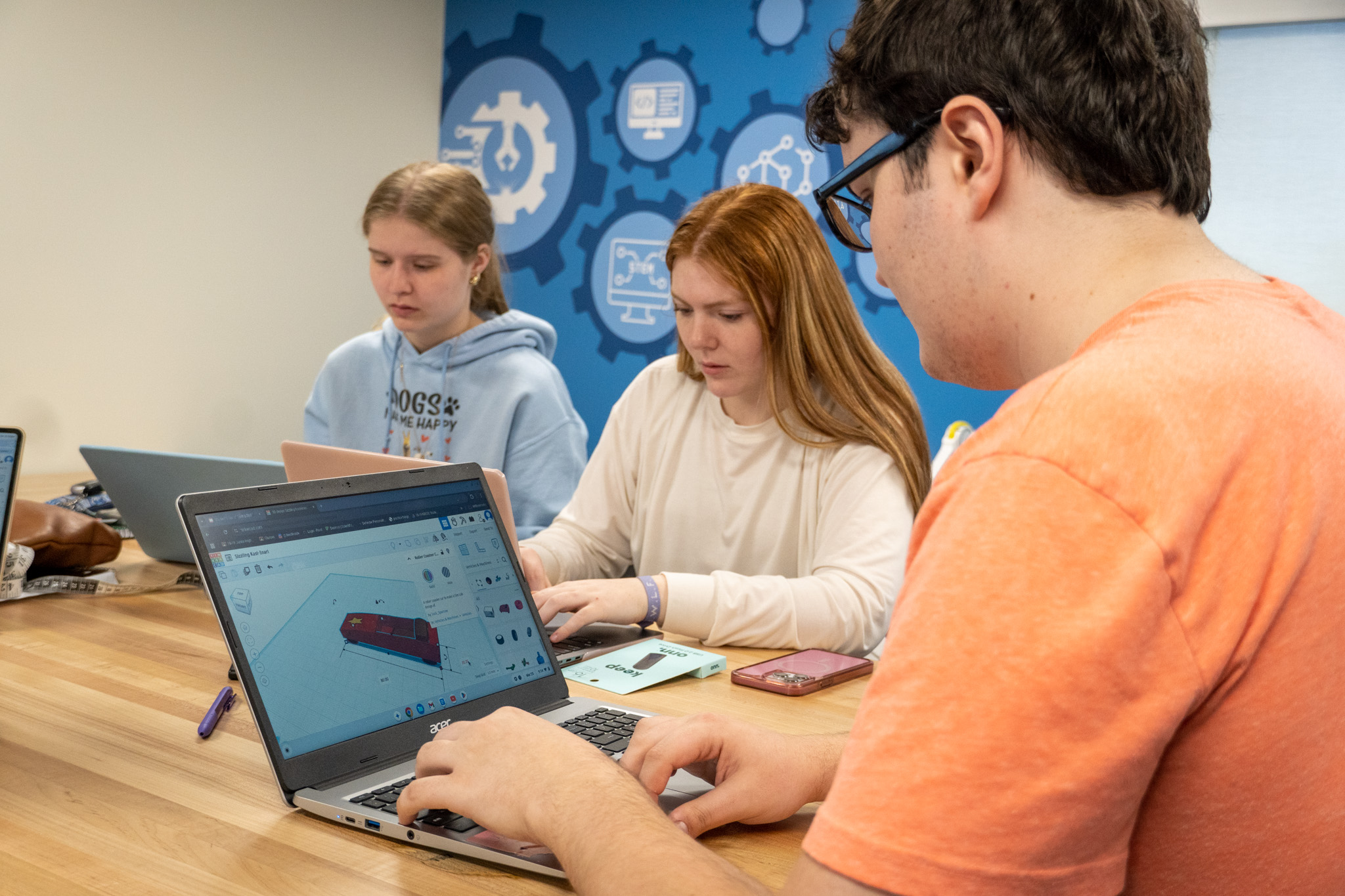 Three young people sit at a long wooden table working on laptops. In the foreground, a student with dark hair and glasses types at an Acer laptop displaying a 3D CAD design of a small vehicle. To his left are two female students also working on laptops. A blue accent wall features gear and icon designs related to STEM fields, suggesting a classroom or educational environment focused on technology and engineering.