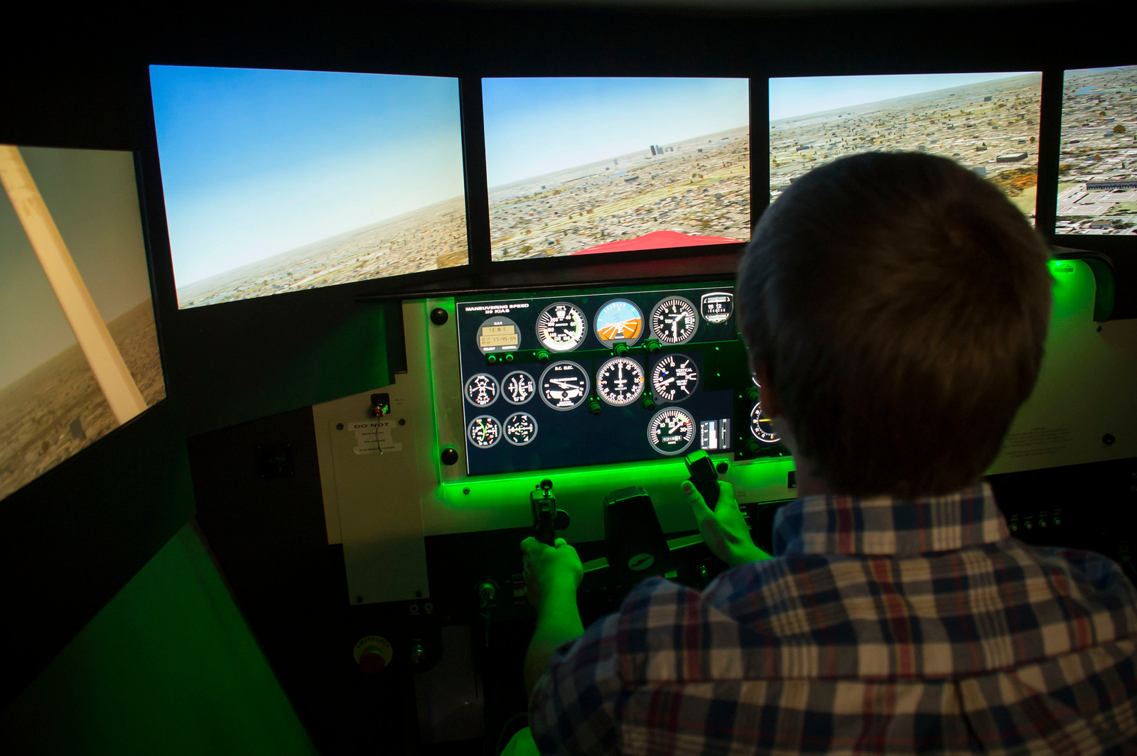 A person in a plaid shirt operates a flight simulator. Multiscreen displays show an aerial view of land, while vibrant cockpit gauges glow green.