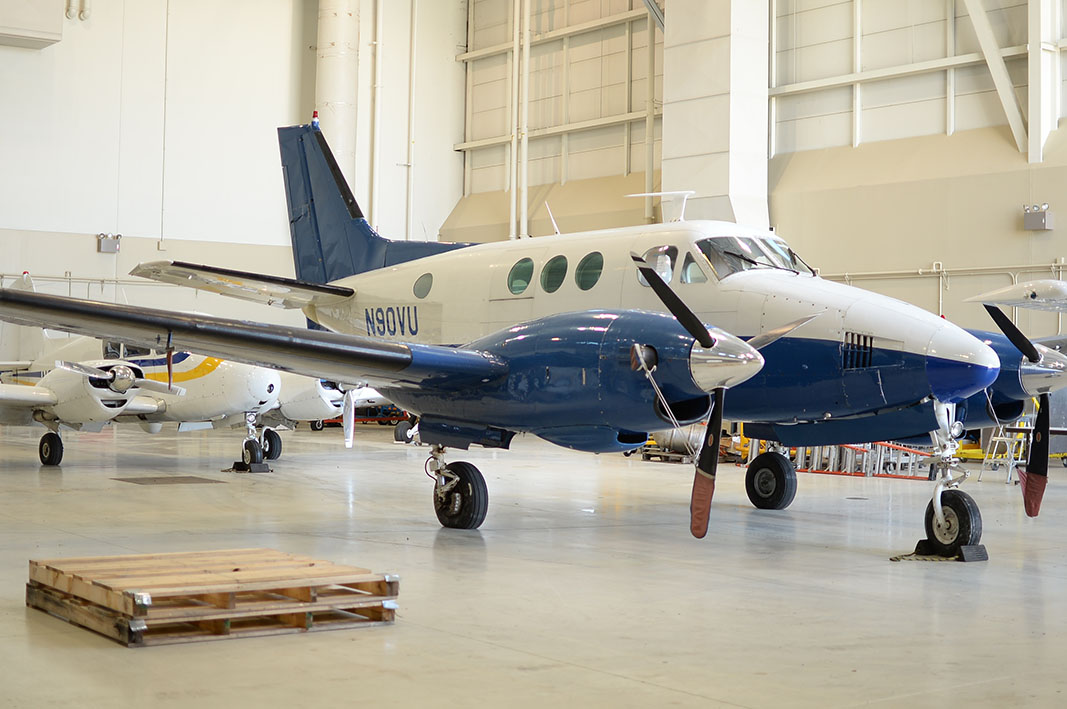 An eye-level shot of a blue and white propeller plane, designated N90VU, parked on a smooth, light-gray floor inside a large aircraft hangar. Additional planes are visible in the background. In the foreground, there’s a wooden pallet.