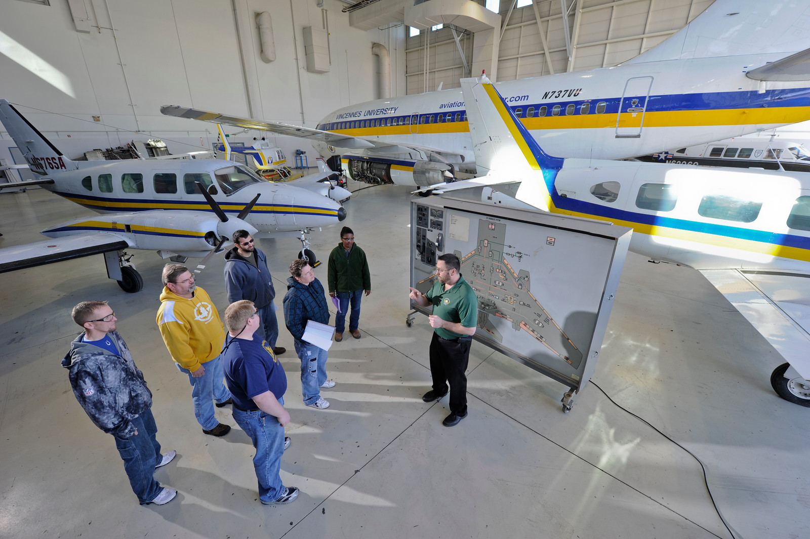Instructor in a hangar explaining using a map, with several planes around. Students listen attentively, creating a focused and educational atmosphere.