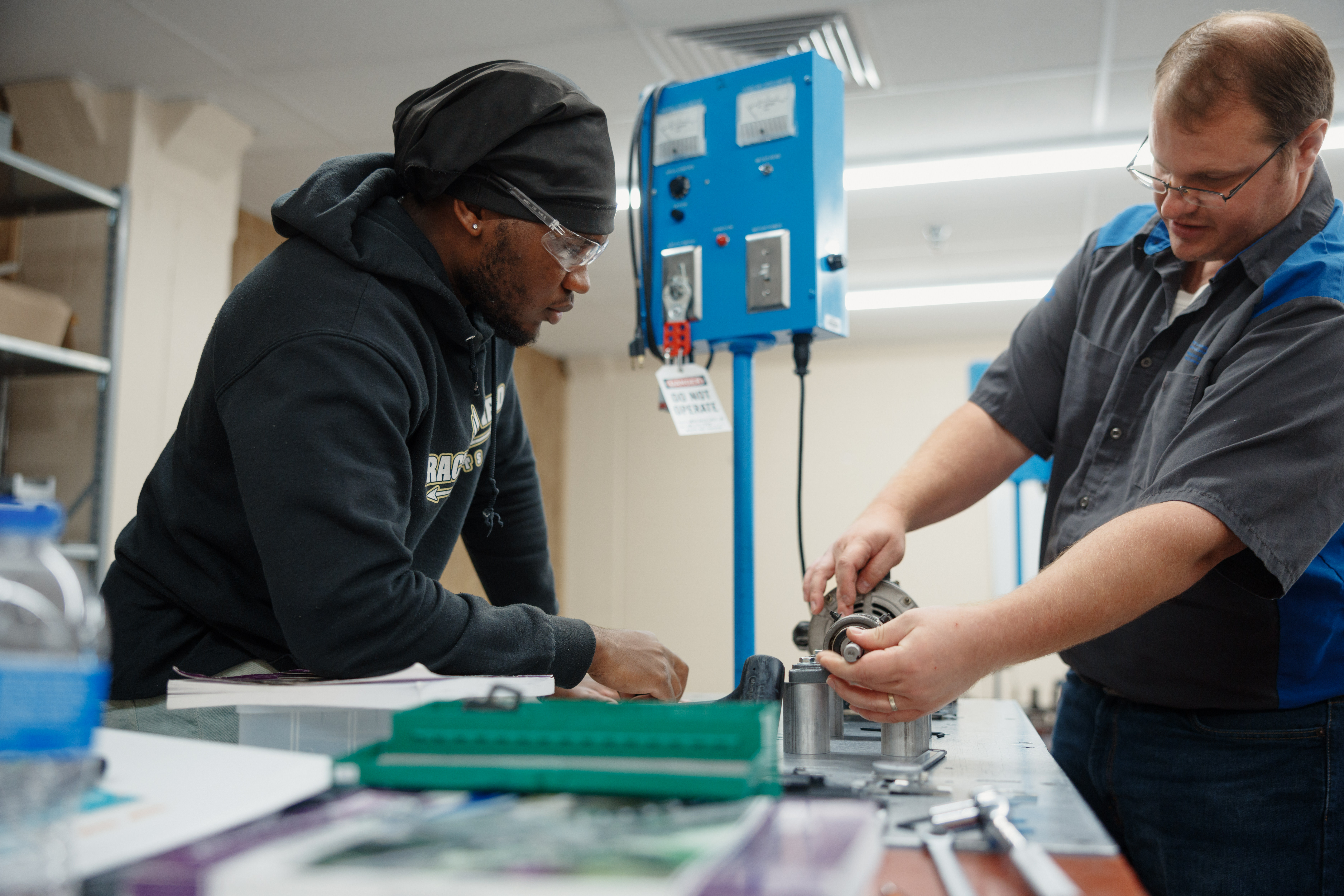 A student and instructor in a workshop discuss machining equipment. One leans over a table observing; the other adjusts equipment. Focused and collaborative atmosphere.