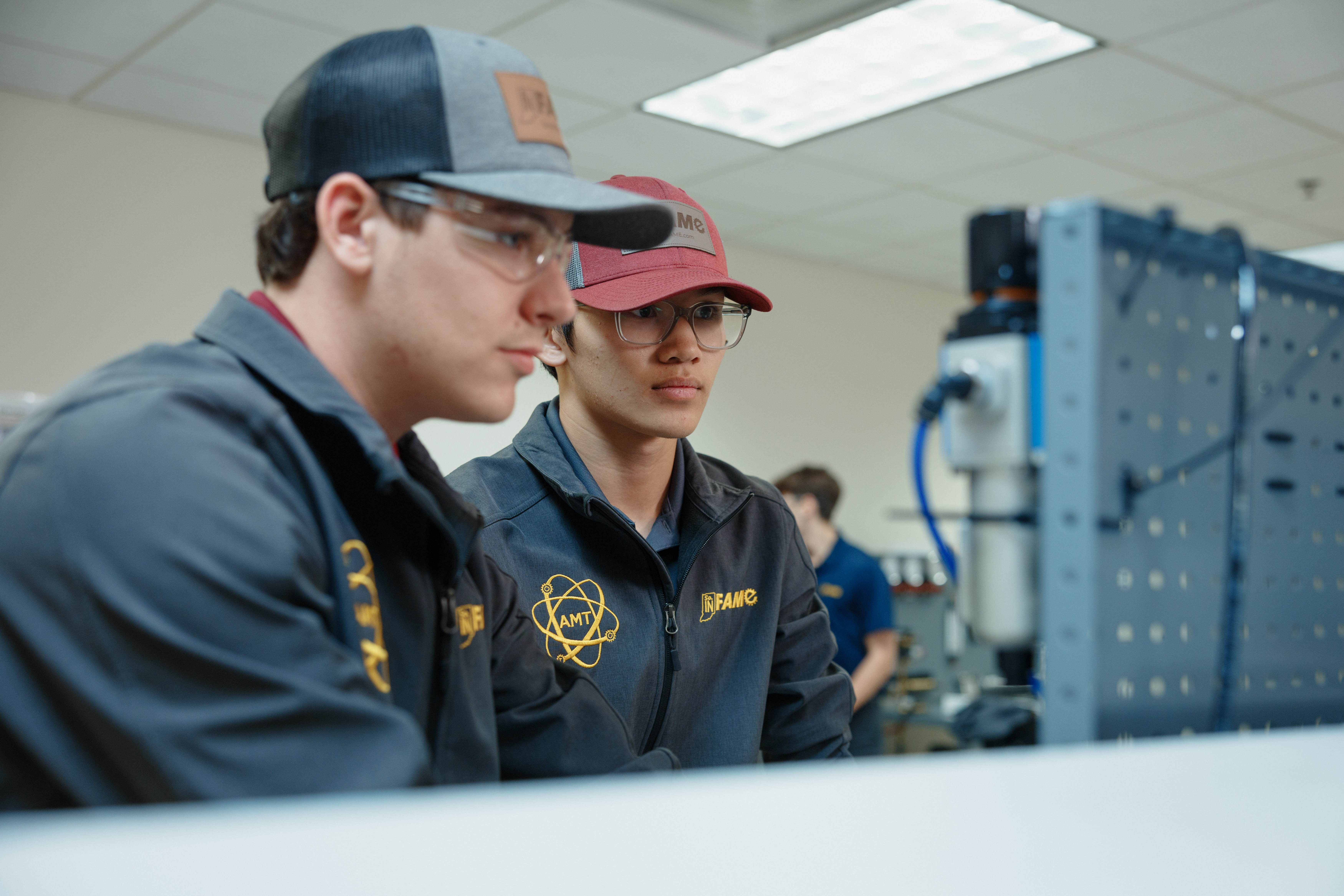 Two young men in hats and protective eyewear focus attentively on a machine in a workshop, wearing jackets with logos. The setting is professional and studious.