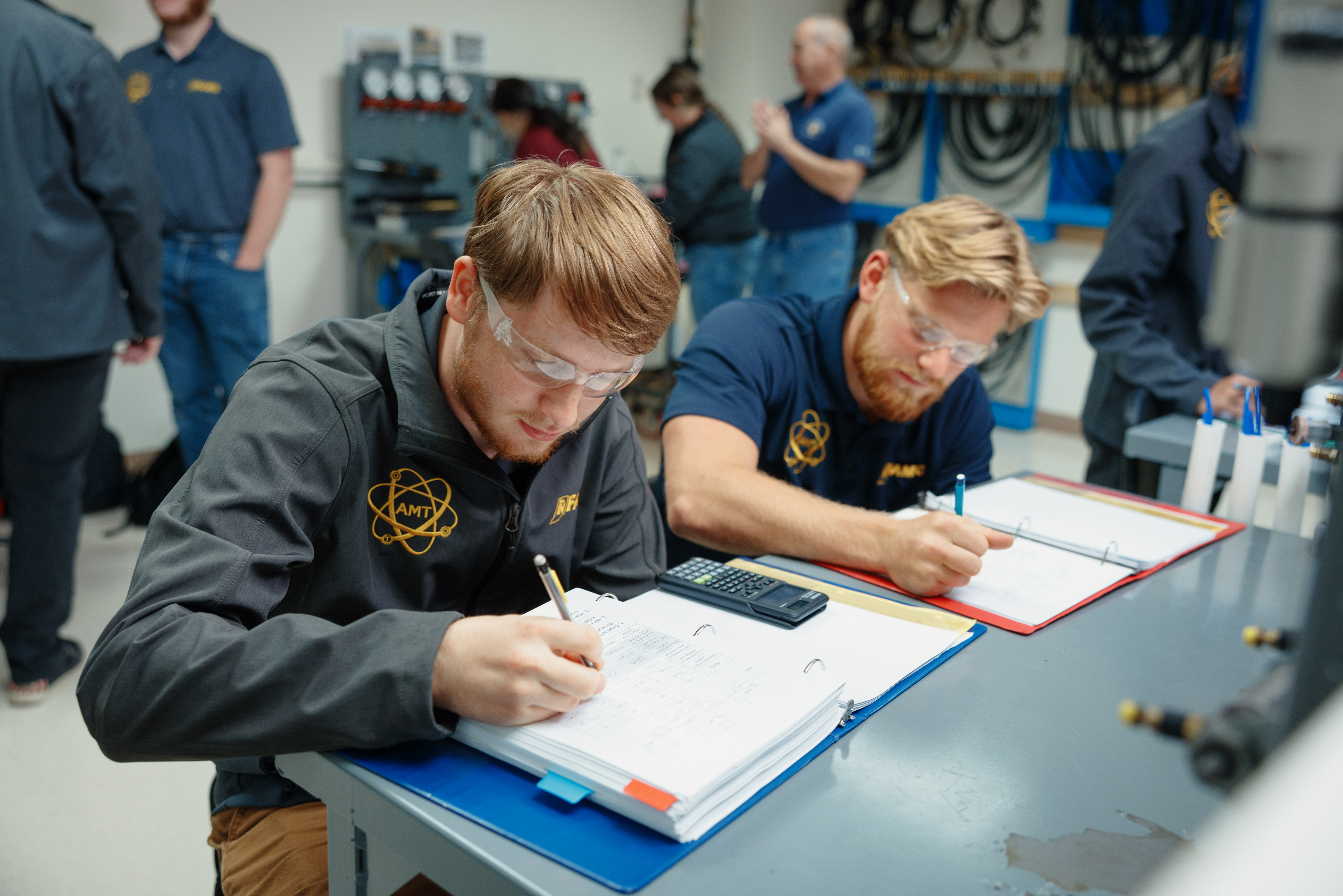 In a workshop setting, two male students wearing safety glasses, each wearing a dark jacket or shirt with a visible "AMT" logo, are diligently taking notes in binders on their respective desks. A calculator sits on one binder. Other students and an instructor are visible in the background, some seemingly engaged in other activities or observing with approval. The atmosphere suggests a hands-on learning environment.