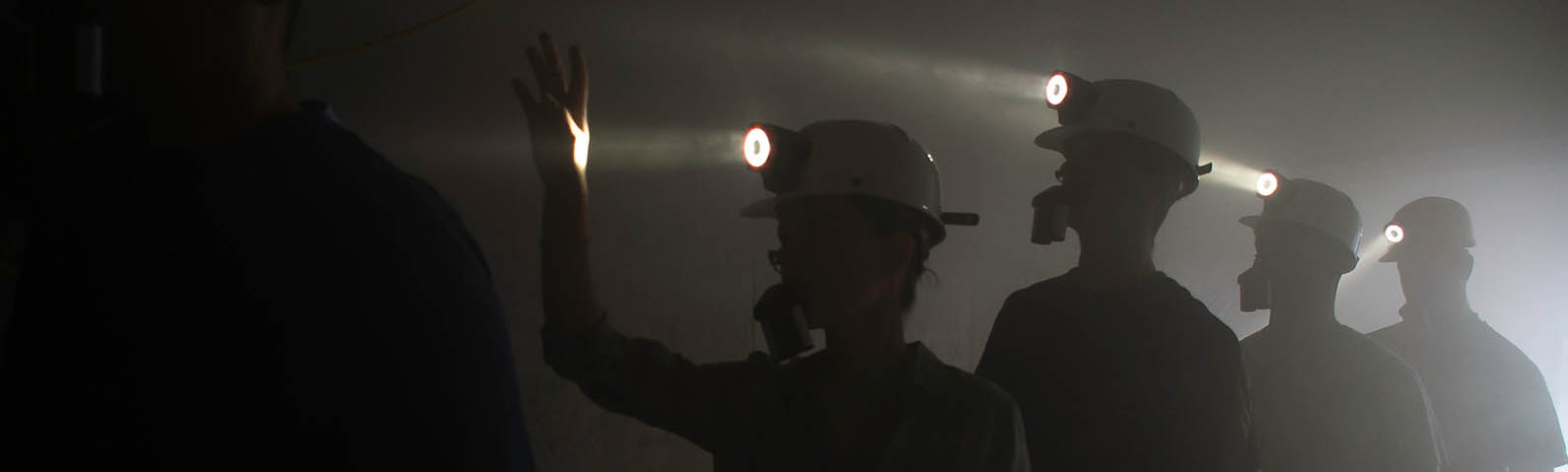 A group of miners are walking through a dark, possibly smoky tunnel. They are wearing protective helmets with headlamps that cast bright beams of light. One miner in the front has a hand raised, possibly signaling or checking conditions.