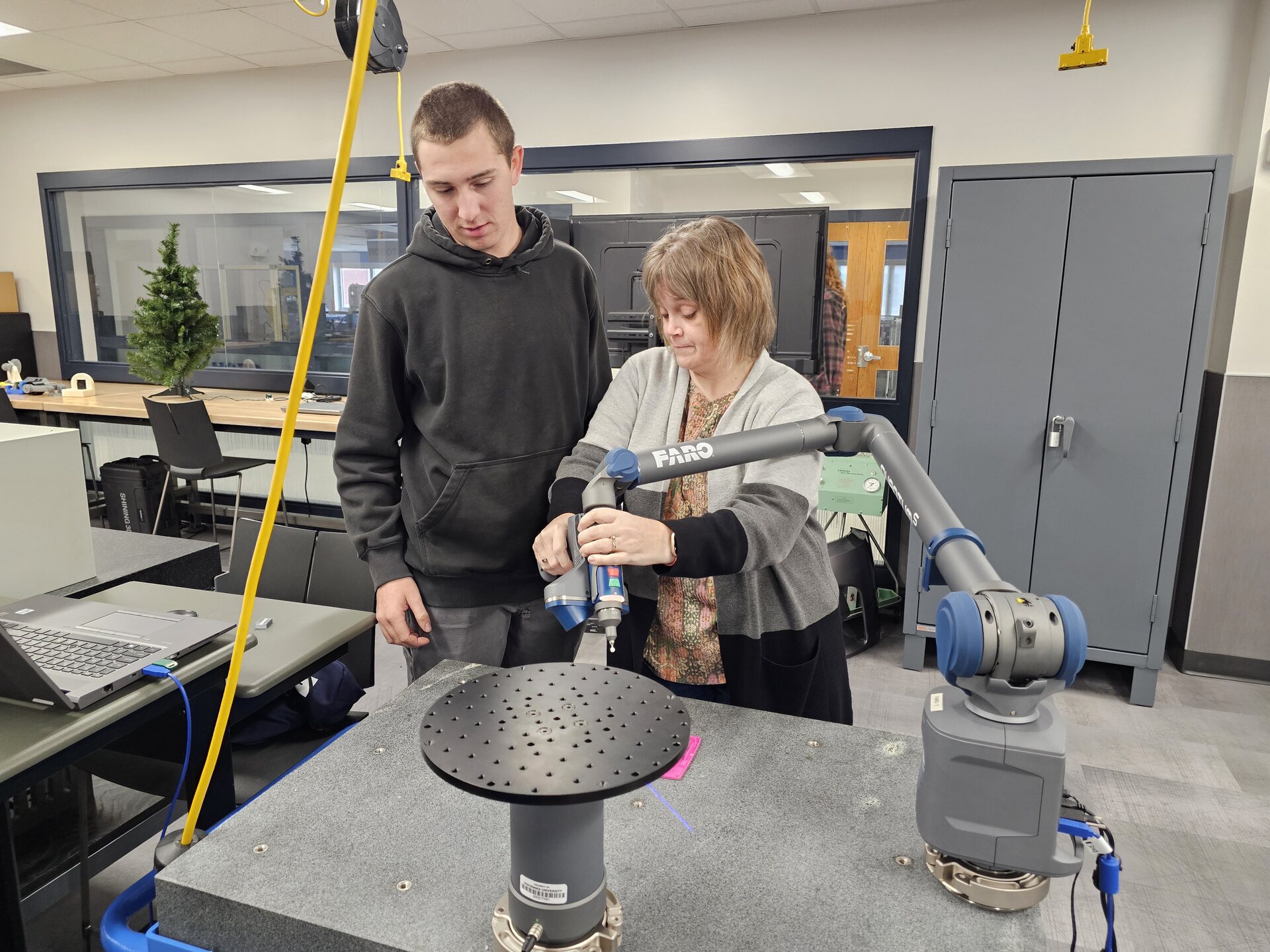 In a well-lit manufacturing lab, a young man in a black hoodie watches as a woman with short brown hair operates a gray articulating-arm coordinate measuring machine, a FARO arm, which is positioned over a circular object mounted on a gray stand. The scene includes various lab equipment, a laptop, an artificial Christmas tree visible through a window, and a gray storage cabinet.
