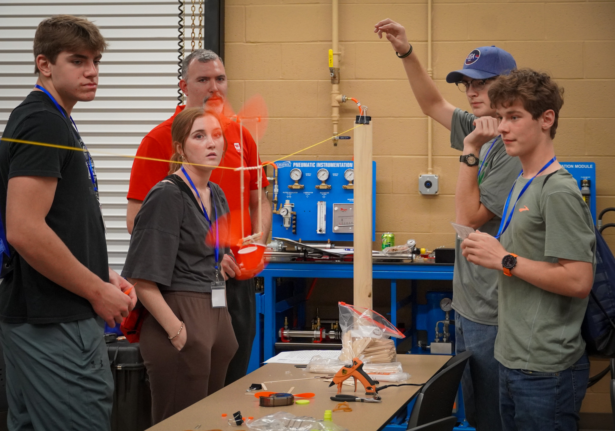 A group of five people, including four students and one instructor, are gathered around a table in a workshop or classroom setting, examining a science project involving a small orange propeller and pneumatic instrumentation. They appear to be intently focused on the project, which is rigged with tubing and attached to a blue Amatrol pneumatic instrumentation module. The students are wearing casual clothing and lanyards. Tools and materials such as glue sticks, craft sticks, and tubing are visible on the table. The instructor, dressed in red, has an orange blur around them perhaps being their moving. The overall atmosphere is one of engaged learning and hands-on experimentation.