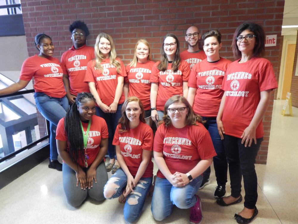 A group of eleven young adults, mostly students, are posing in a hallway inside of a building. A brick wall is behind them and tile is beneath them. All are wearing matching red t-shirts with "Vincennes" and either "Psychology" or "Social Work" printed on them. Some are standing while some are kneeling. Most are smiling to the camera.