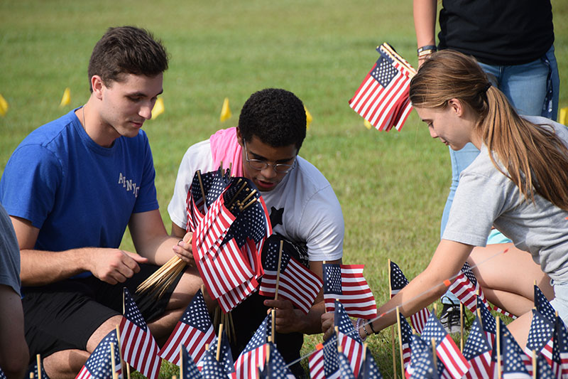 Three young volunteers kneel on a grassy field, placing small American flags into the ground. Many other flags have already been planted. The volunteers, two men and one woman, are dressed casually. A yellow ribbon marks the background behind the flag display.