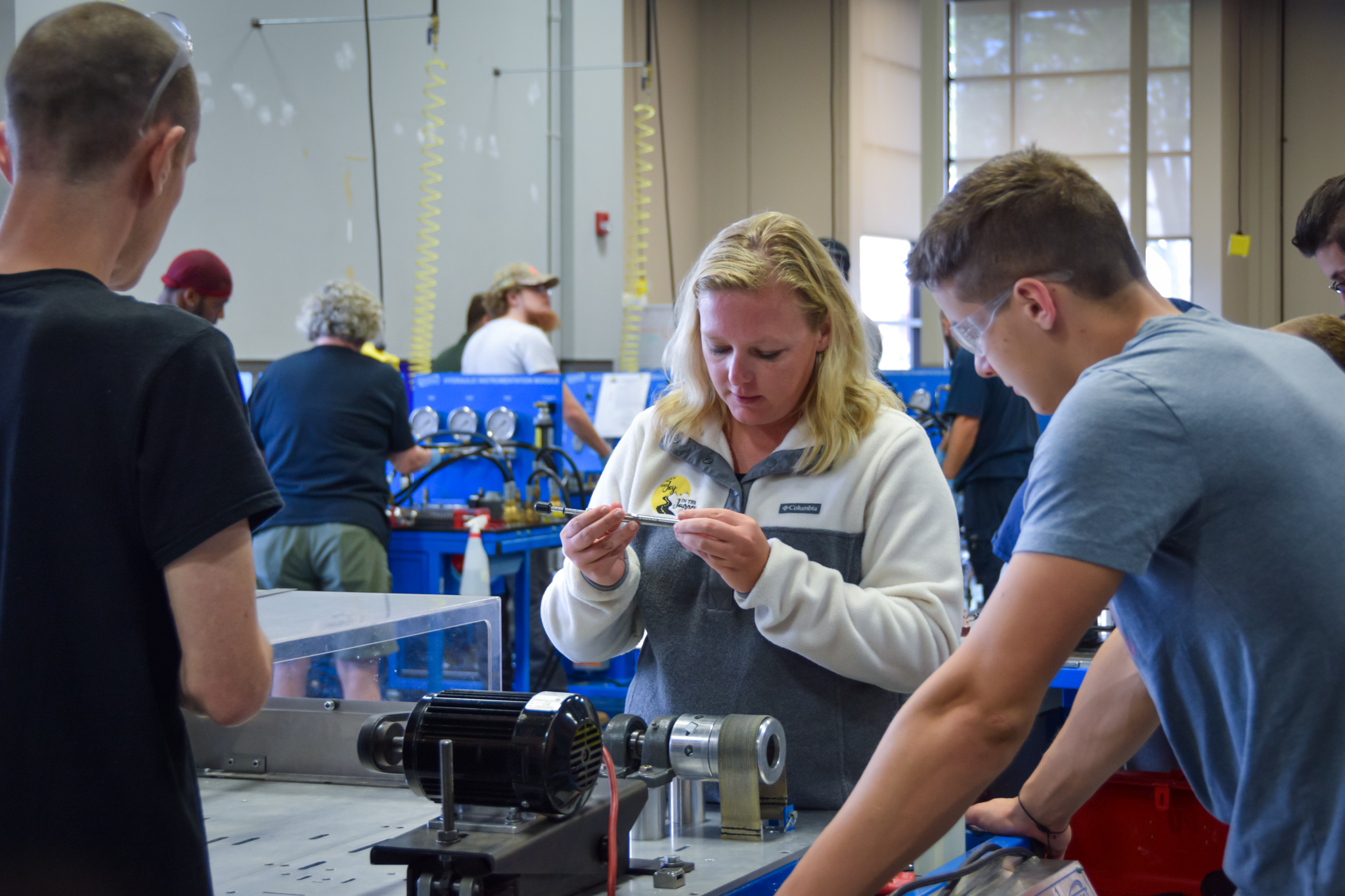 In a brightly lit workshop, several students are engrossed in a hands-on mechanical project. A woman with blonde hair, wearing a fleece jacket, carefully examines a small metal component in her hands. Around her, other students are clustered around workbenches with various tools and machinery, including motor and pulleys. The scene conveys a learning environment focused on practical skills.