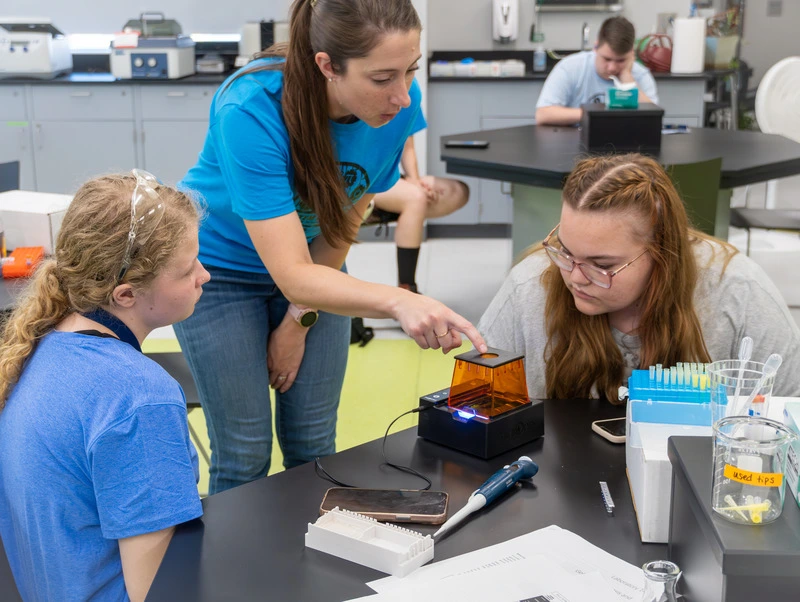 A teacher and two students in a lab engage with a small device on a table.