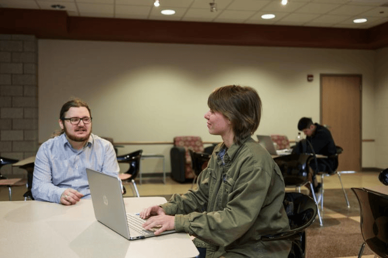 Two people sit at a table with a laptop in a casual, well-lit room.