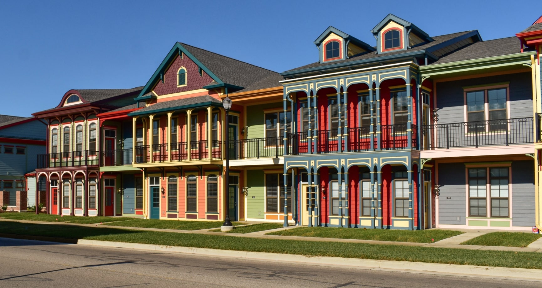 A row of multi-story, Victorian-style townhouses stands side-by-side, each painted in a different vibrant color scheme. The houses face the street, each has its own small, grassy yard. Many houses are decorated with a columned porch or balcony. The houses are under a clear, deep blue sky.