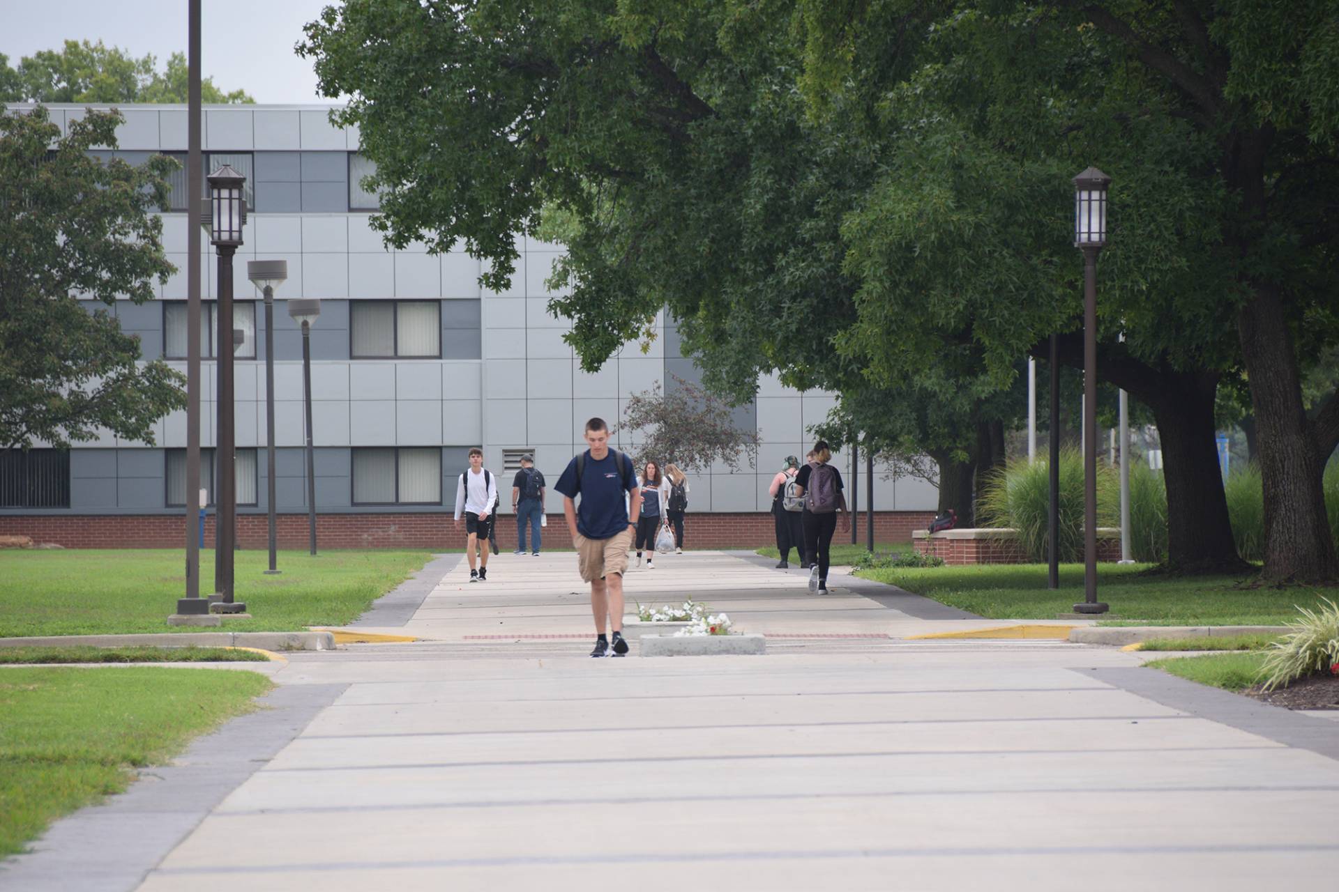 A wide, paved pathway cuts through a college campus. Students are walking along the path in both directions. In the foreground, a young man in a navy t-shirt and khaki shorts walks toward the viewer. Behind him are other students, some carrying backpacks; to the left and right are grassy areas with trees and lamp posts. A modern, multi-story building with gray and white panels serves as a backdrop. The sky is overcast, casting a soft light on the scene.