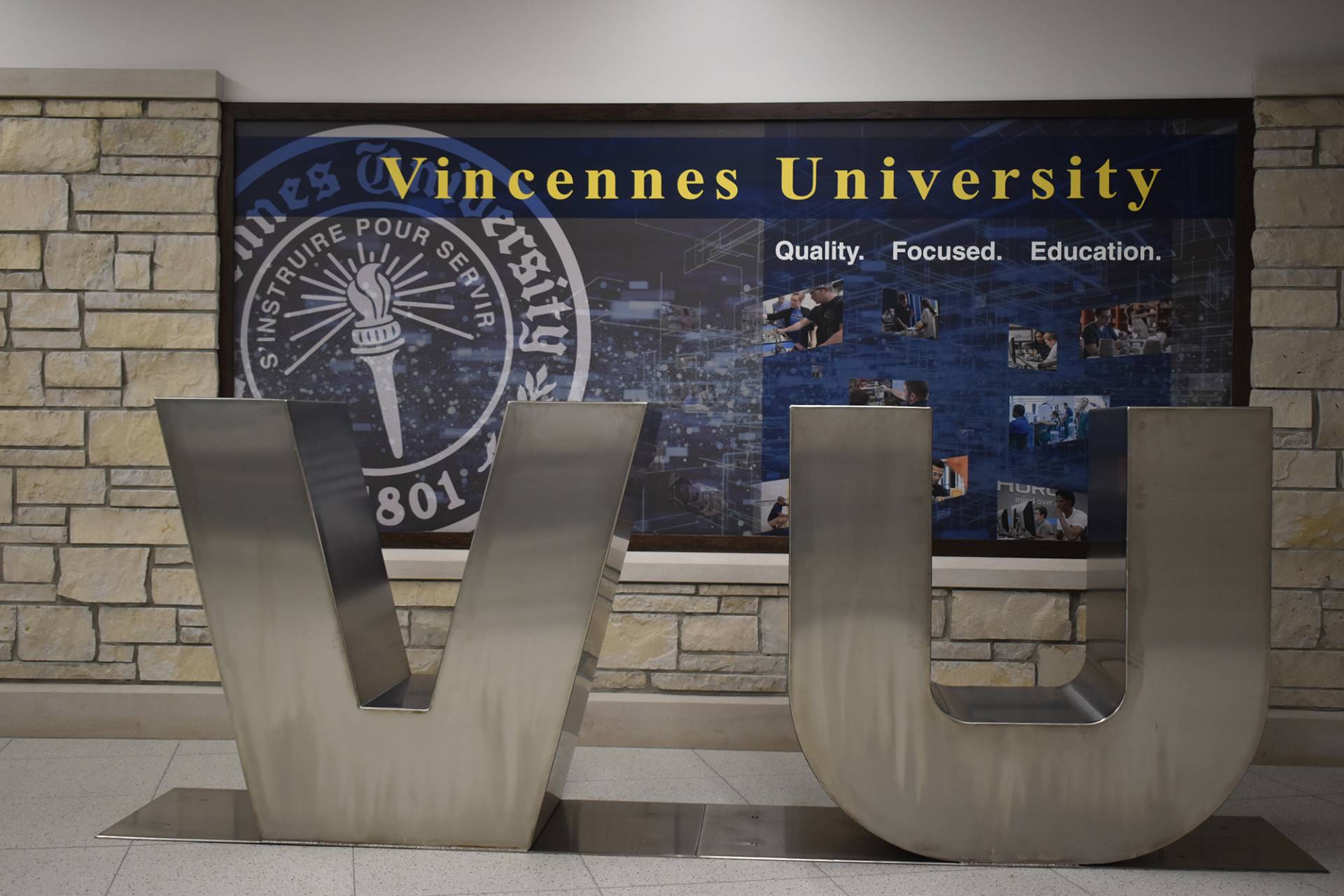 A photograph of the Vincennes University campus, featuring a large, brushed metal sculpture of the letters "VU" in the foreground. Behind the sculpture is a wall display with the university's name, "Vincennes University," emblazoned at the top in bold yellow letters. Below the name are the words "Quality. Focused. Education.", and further below are smaller images depicting students and faculty engaged in various activities. To both sides of the display are walls constructed from light brown stone blocks. The sculpture is on a speckled light-colored floor.