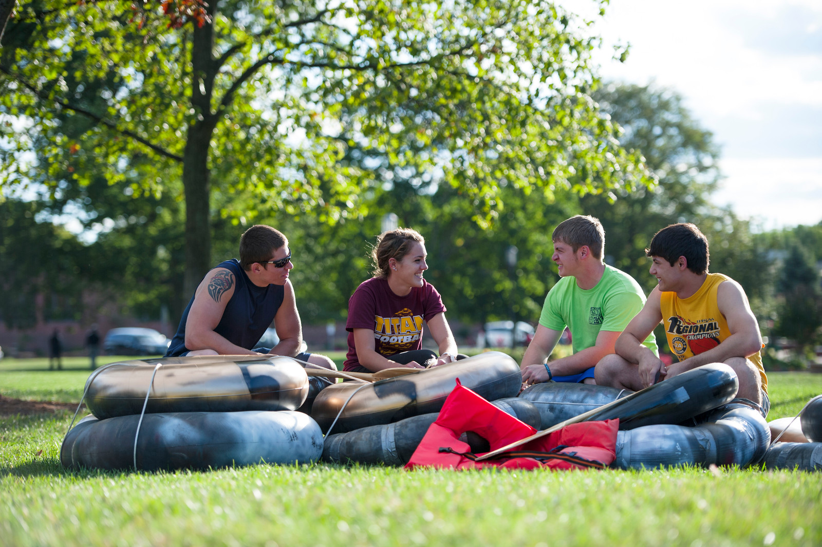 Four college-aged students are sitting on a makeshift raft made of interconnected inner tubes, on a grassy field under the shade of a large tree. They are all smiling and engaged in conversation. The raft is surrounded by several red life jackets and wooden paddles. The students are dressed in casual clothing; one wears sunglasses and has a tattoo visible on their arm, and two of them wear shirts with college team logos. In the background, there are trees and blurred figures. The overall atmosphere is relaxed and indicates a campus or outdoor activity.