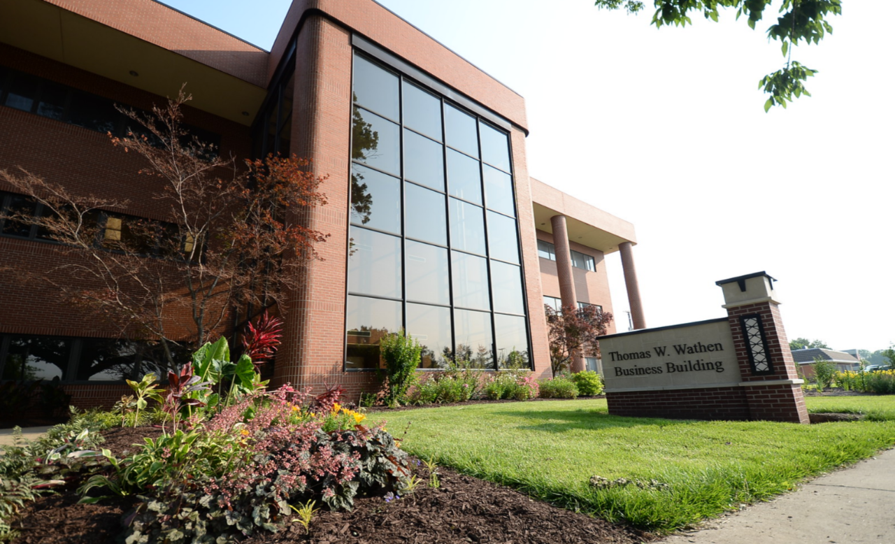 Exterior shot of the Thomas W. Wathen Business Building. The building has a red brick facade with a modern design featuring large, grid-patterned windows. A well-maintained lawn and a flower bed with various colorful plants are at the building's base. The building's sign sits nearby on the right side.