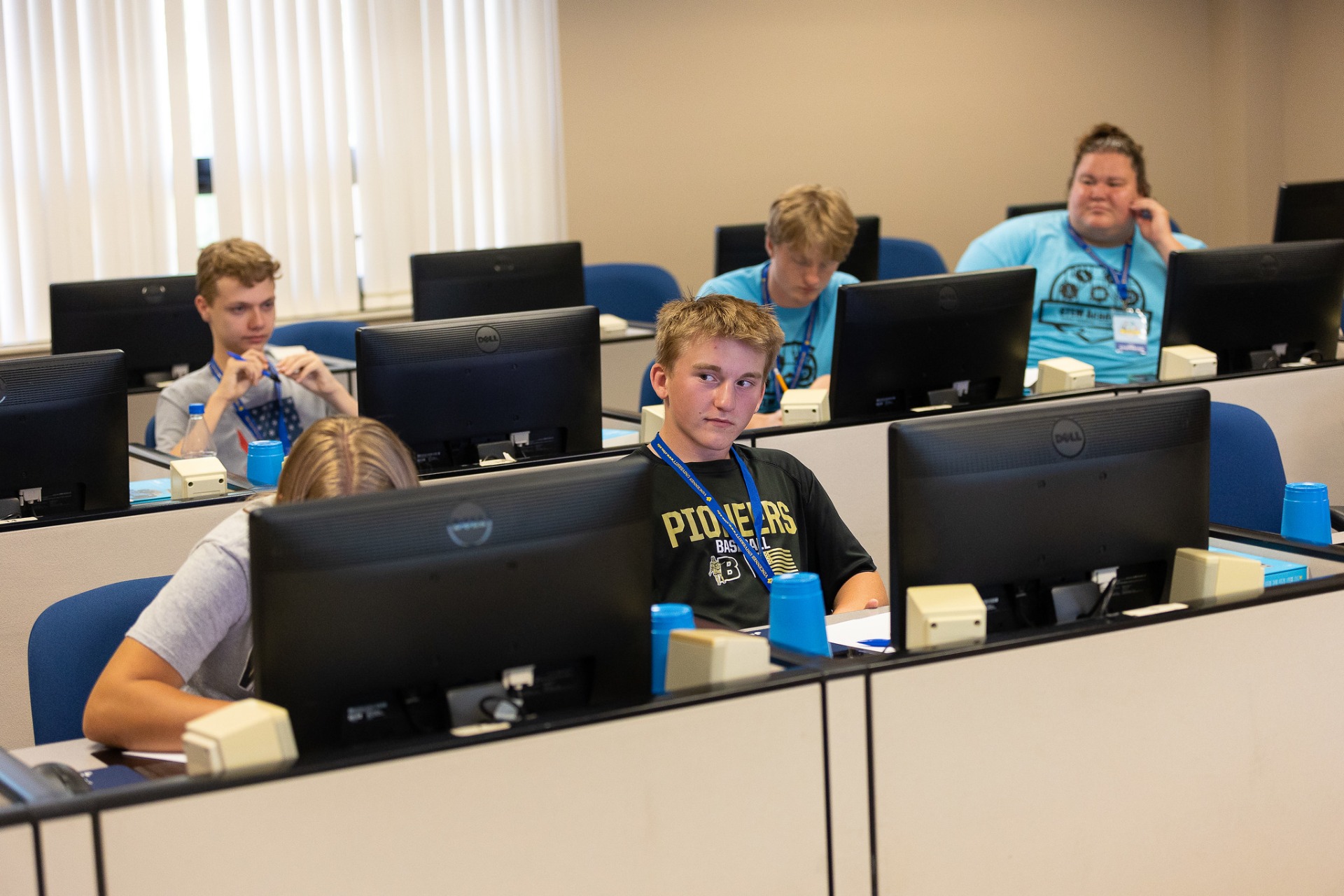 A classroom scene with students sitting at individual computer workstations. The students, all young adults, are focused on their monitors. Each workstation features a Dell computer, a blue cup, and a small cream-colored electrical box. The students are wearing lanyards around their necks, suggesting a specific event or course. The room has light-colored walls and window blinds, indicating a formal learning environment.